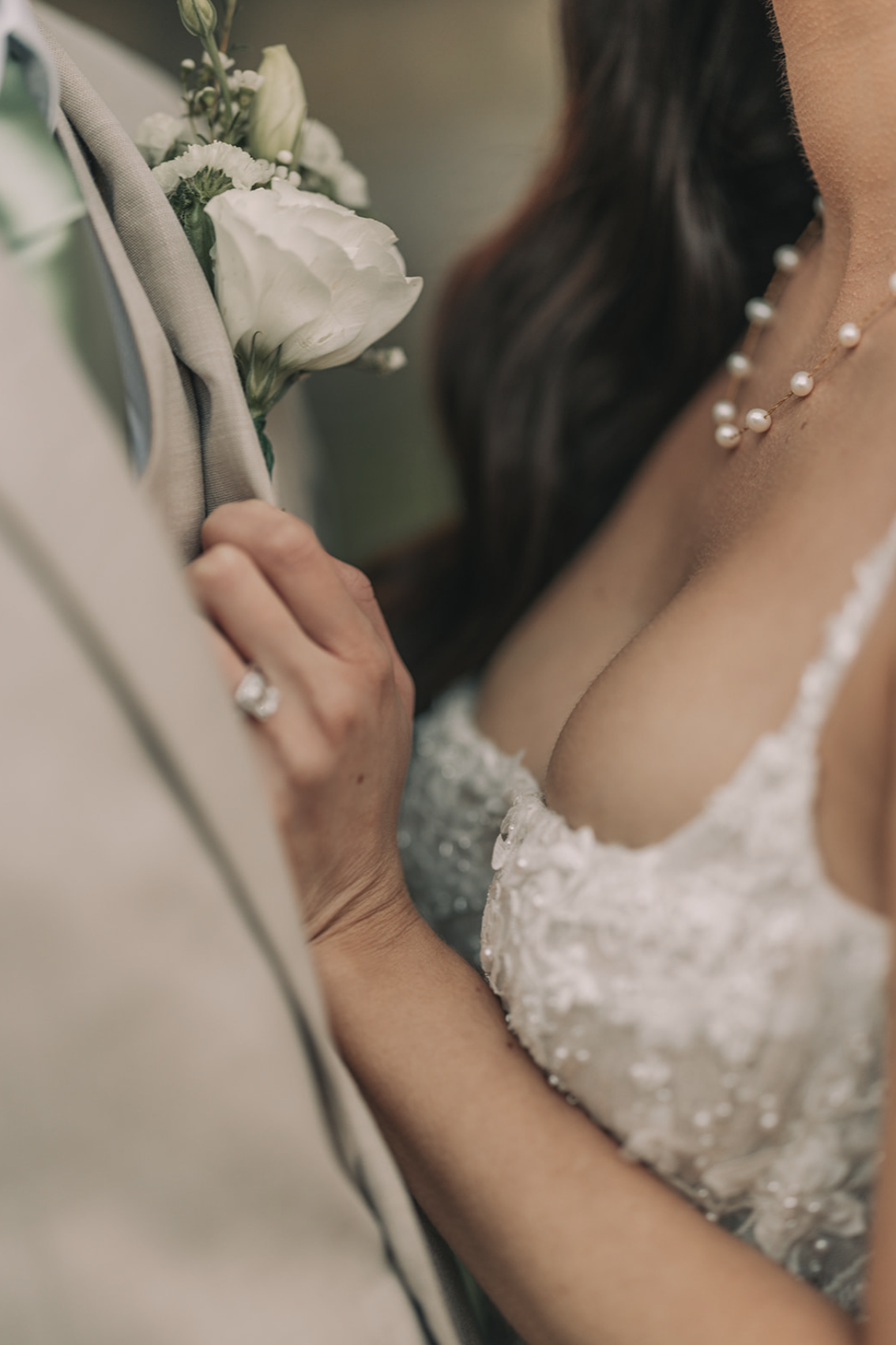 Close-up of a bride and groom during a wedding, with focus on the bride's hand and wedding ring, the groom's boutonniere, and the bride's pearl necklace and wedding dress.