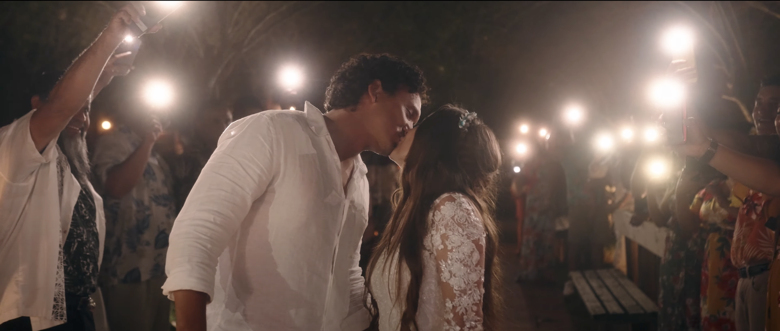 A couple shares a kiss during their wedding reception, surrounded by friends and family with string lights overhead in an outdoor setting.