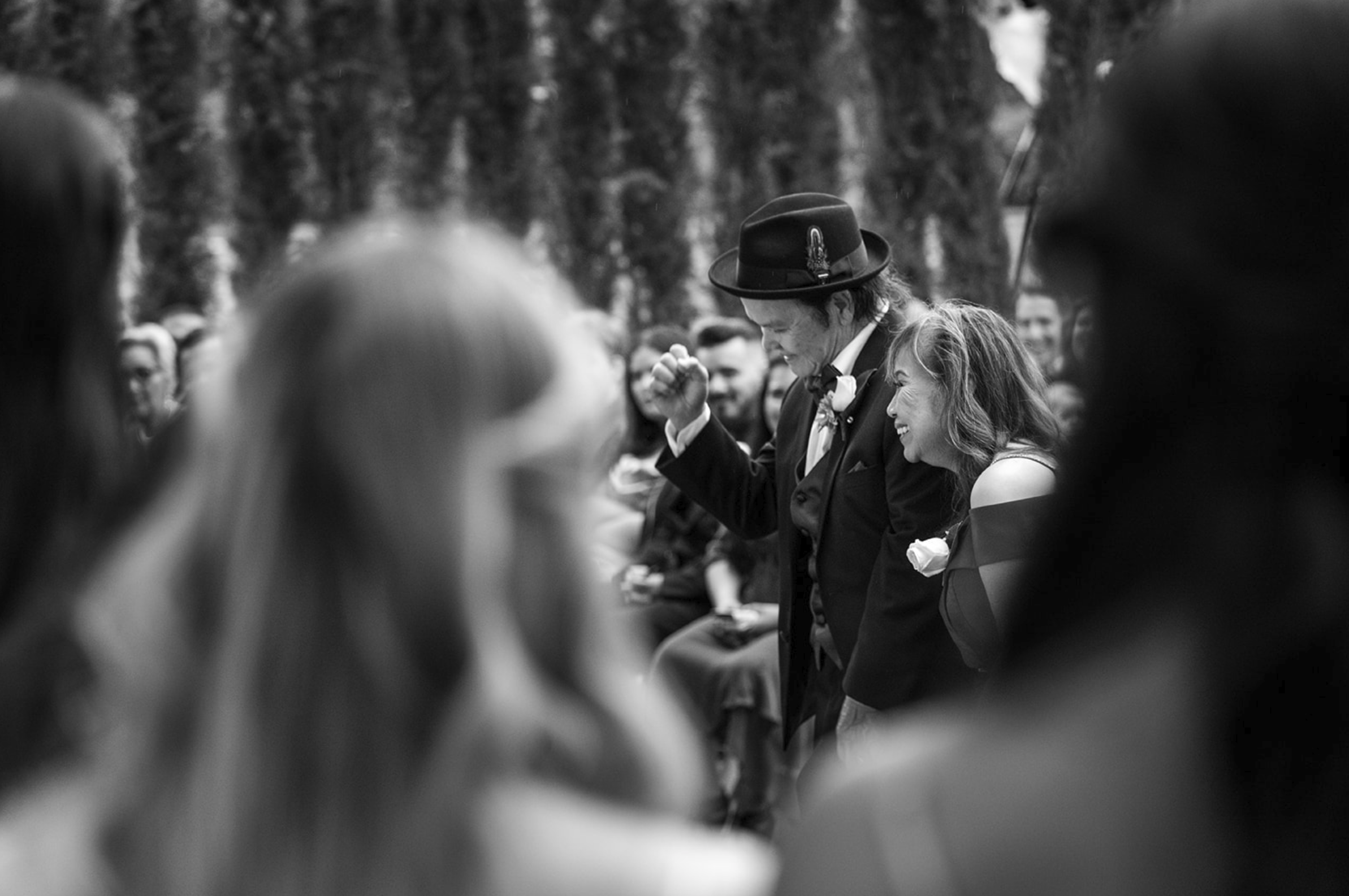 Black and white photo of wedding ceremony outdoors, with a man in a suit and hat raising his fist in celebration, surrounded by smiling guests.