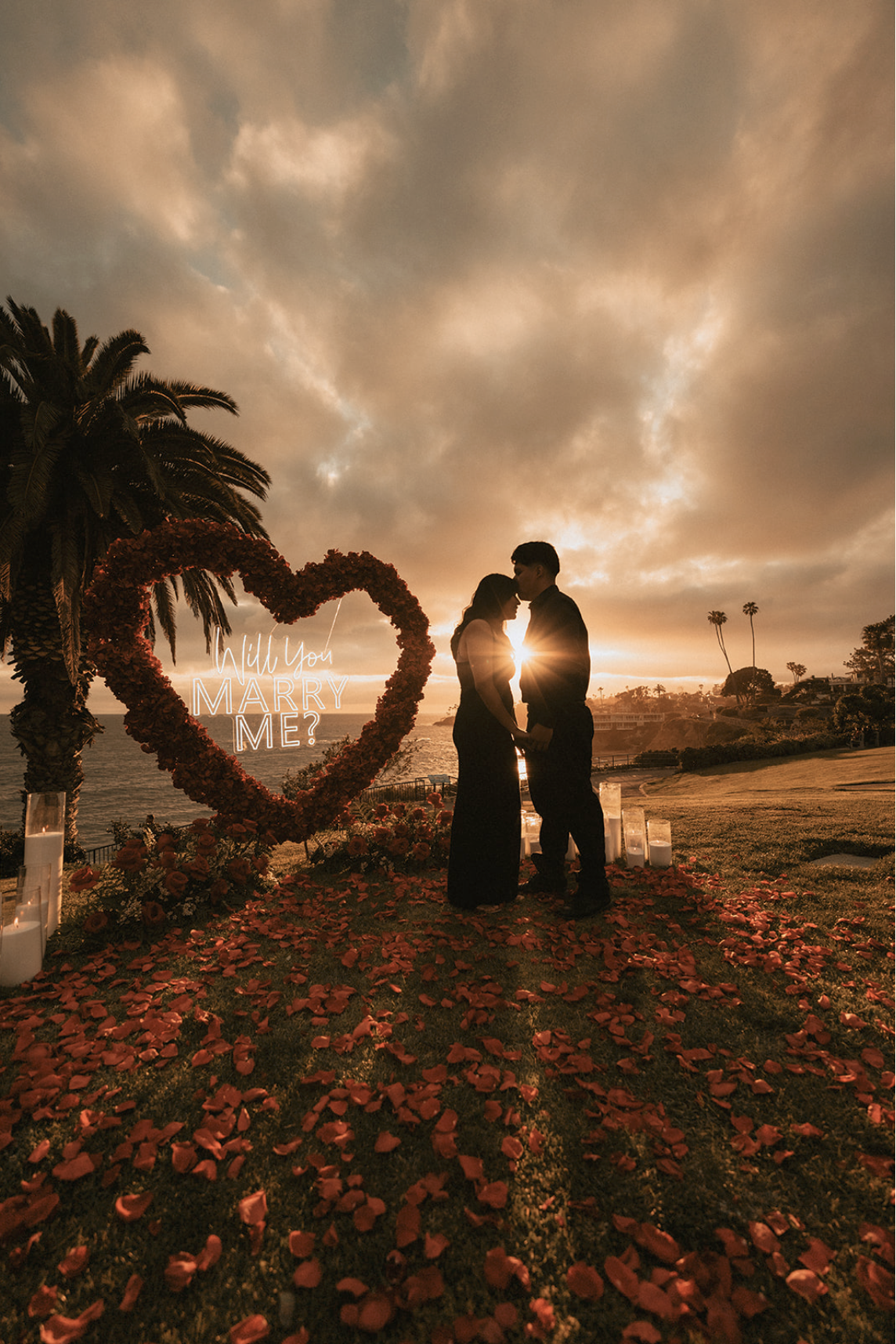 A couple standing close together at sunset during a marriage proposal, surrounded by rose petals and candles, with a heart-shaped floral arrangement that says 'Will You Marry Me?'