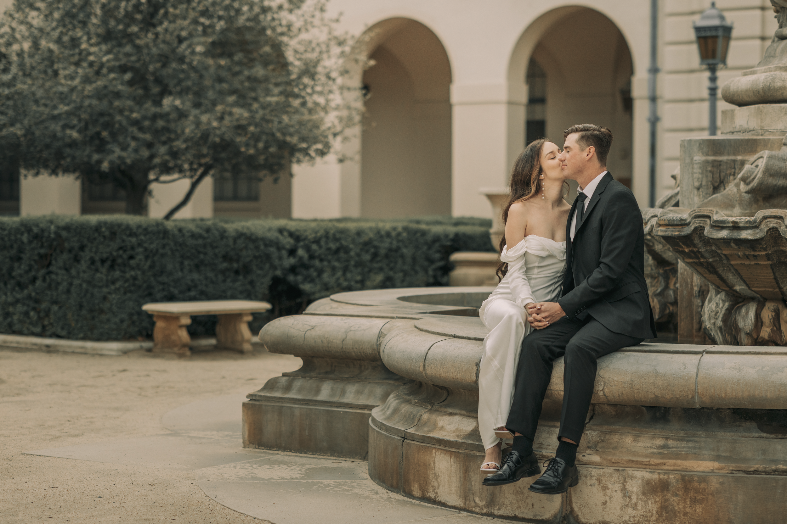 A couple in wedding attire sharing a kiss while sitting on the edge of a stone fountain in an outdoor courtyard with greenery and historic architecture in the background. Pasadena City Hall