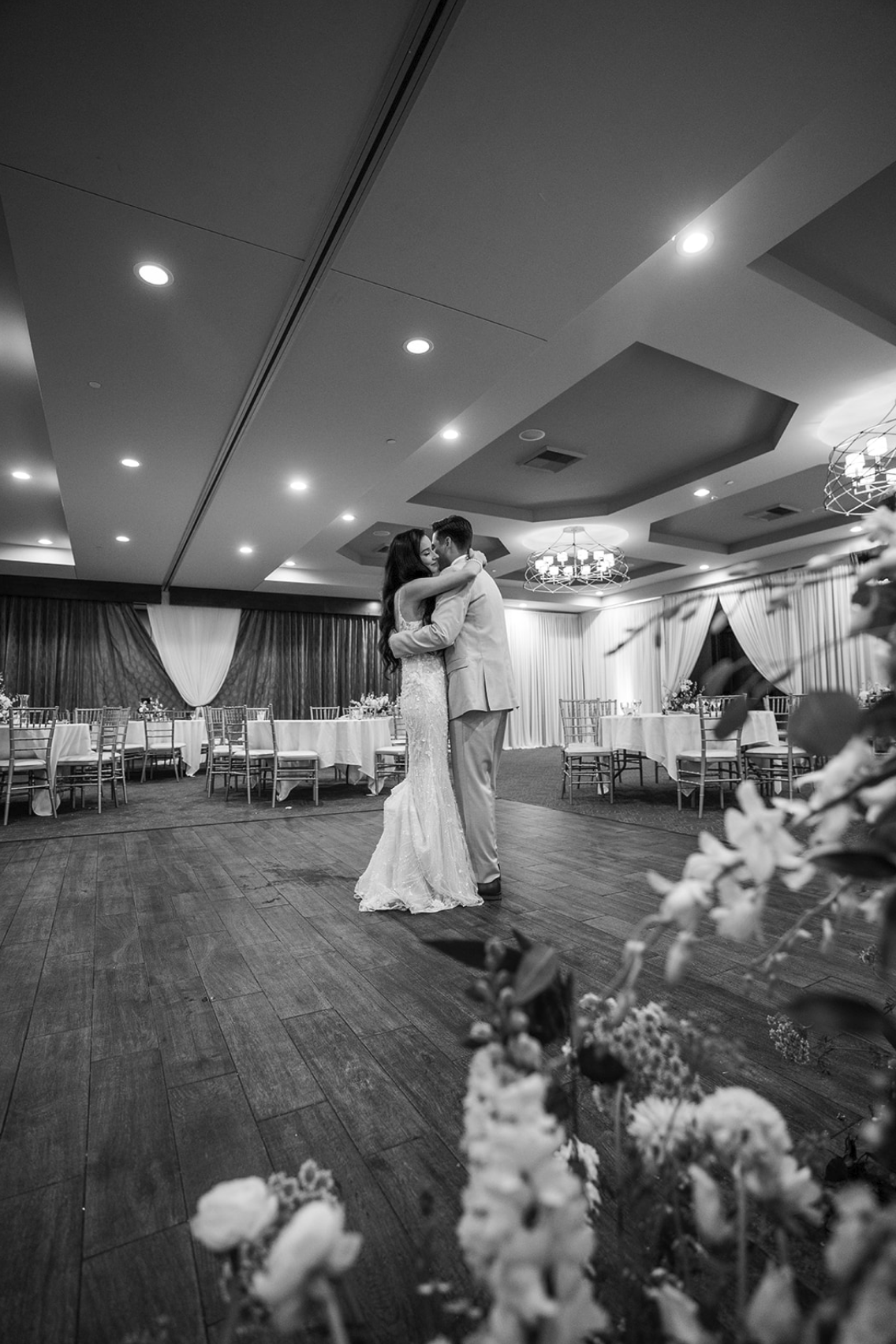 A black and white photo of a newlywed couple hugging and dancing together in an elegant indoor reception hall, surrounded by decorated tables and chairs. Wedgewood wedding venue Chino Hills.