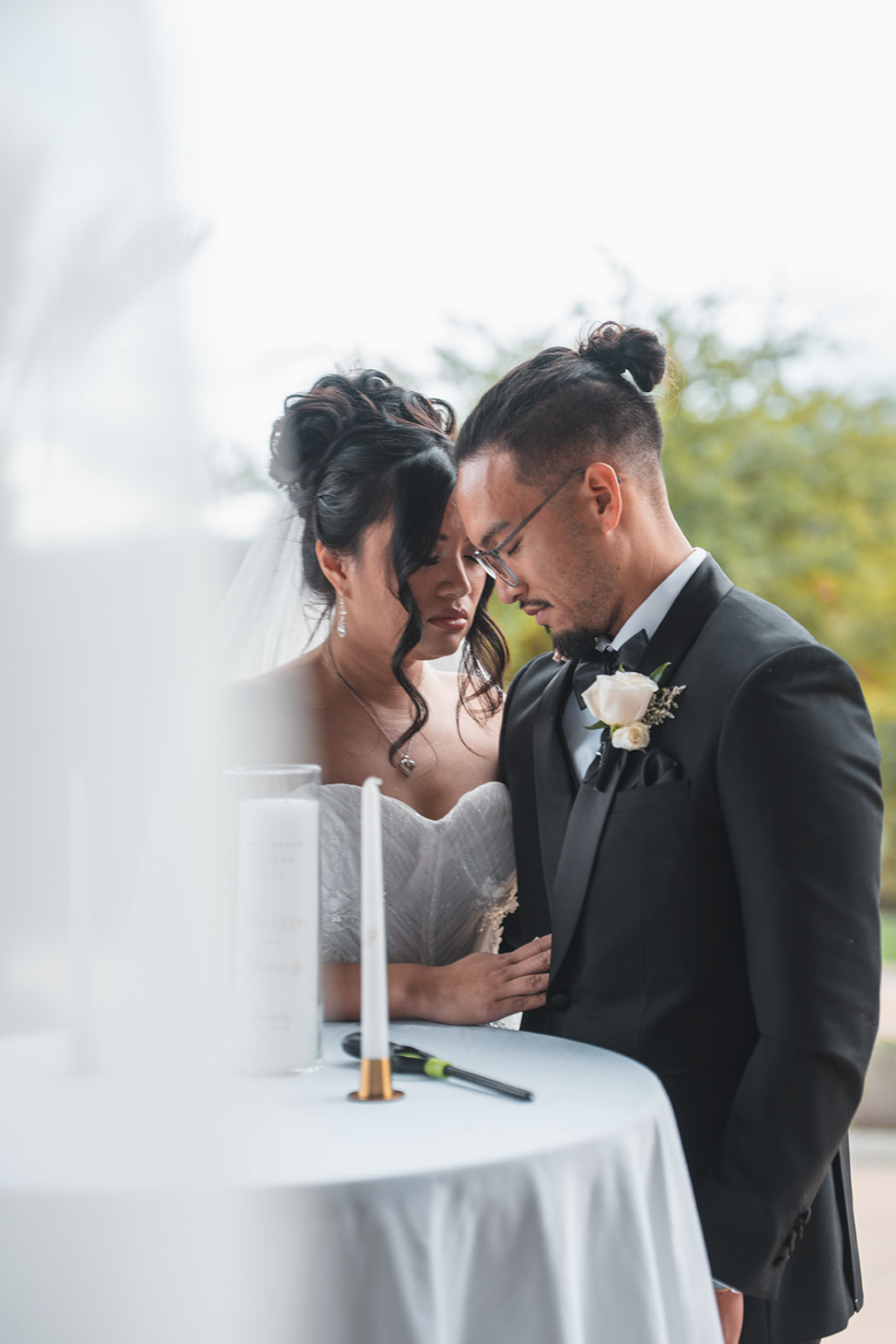A bride and groom with heads bowed and eyes closed during their wedding ceremony outdoors.