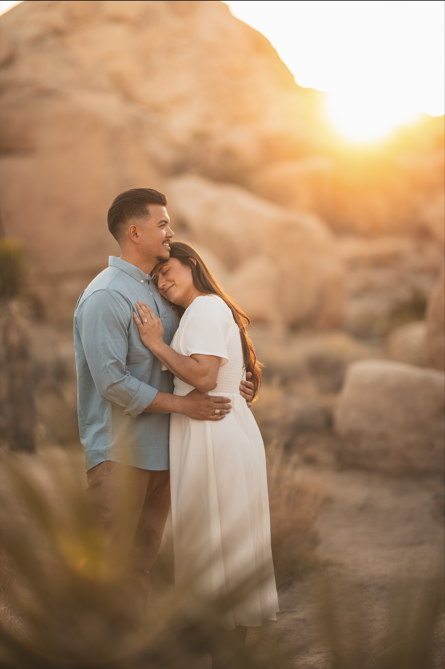 A couple embraces in a rocky outdoor setting during sunset, smiling and content.