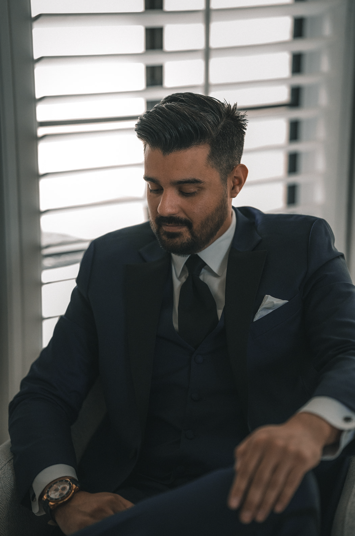 Man in a dark blue suit and tie sitting near window shutters, looking downward with his eyes closed.
