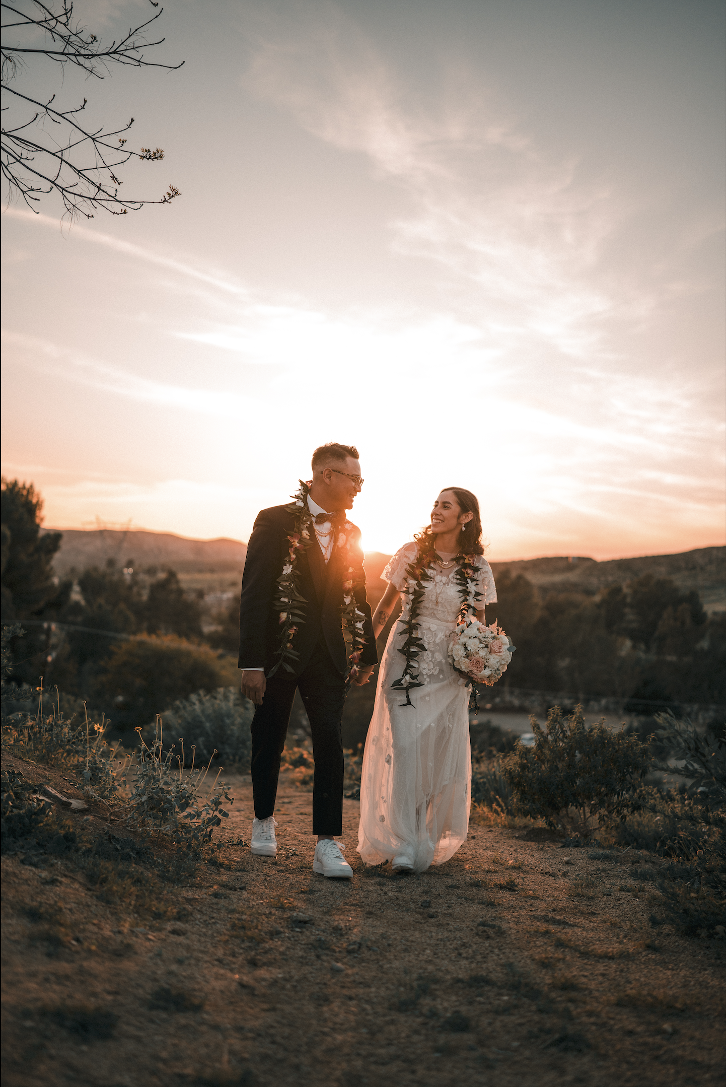 A bride and groom walking hand in hand on a dirt path during sunset, smiling at each other, with the bride holding a bouquet of flowers. The groom is wearing a suit and glasses, and both are wearing leis.