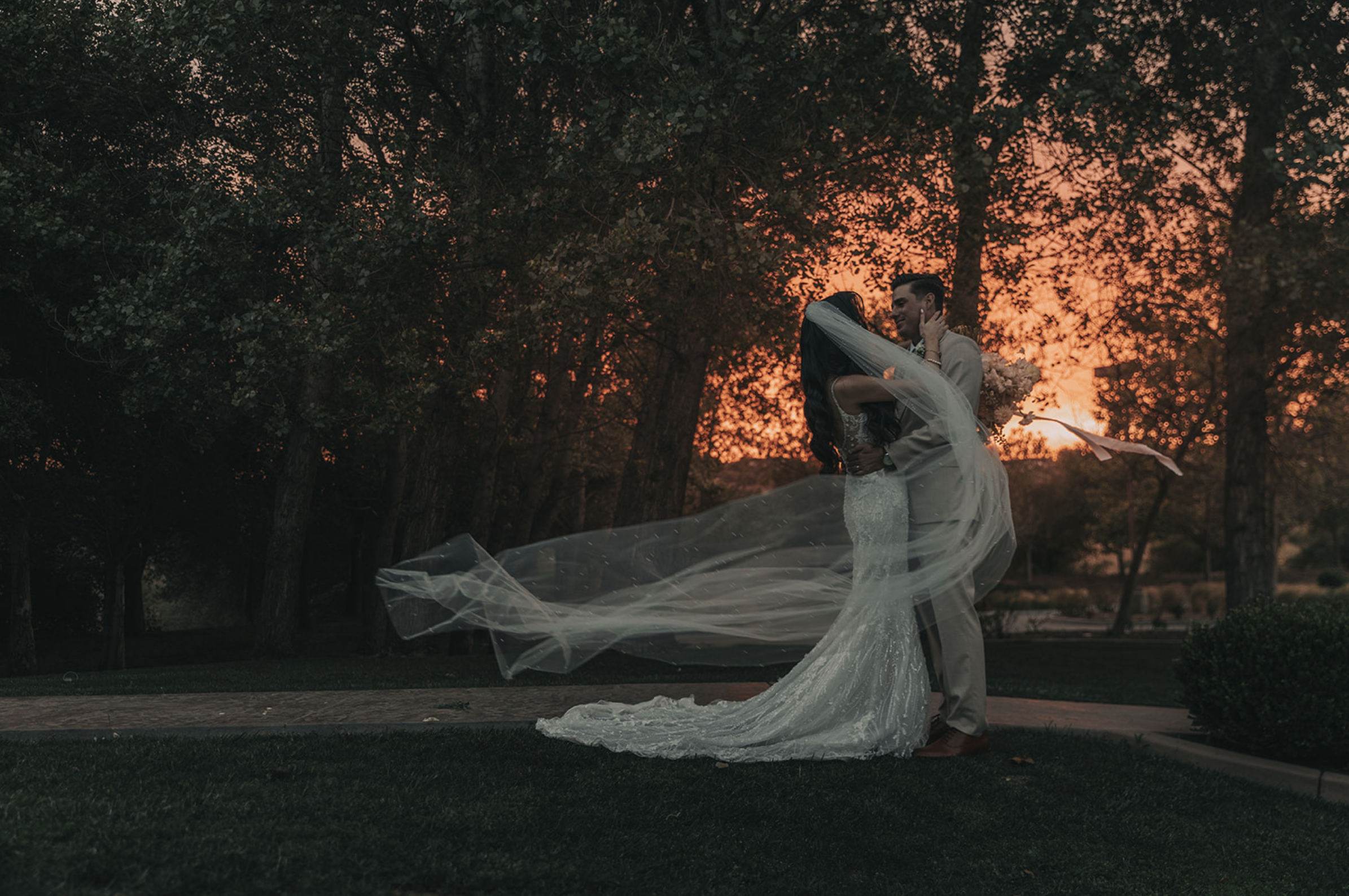 A newlywed couple embracing and kissing outdoors during moody sunset, with the bride in a white wedding gown and veil, and the groom in a light-colored suit, surrounded by trees and a sunset sky. Wedgewood wedding venue Chino Hills.