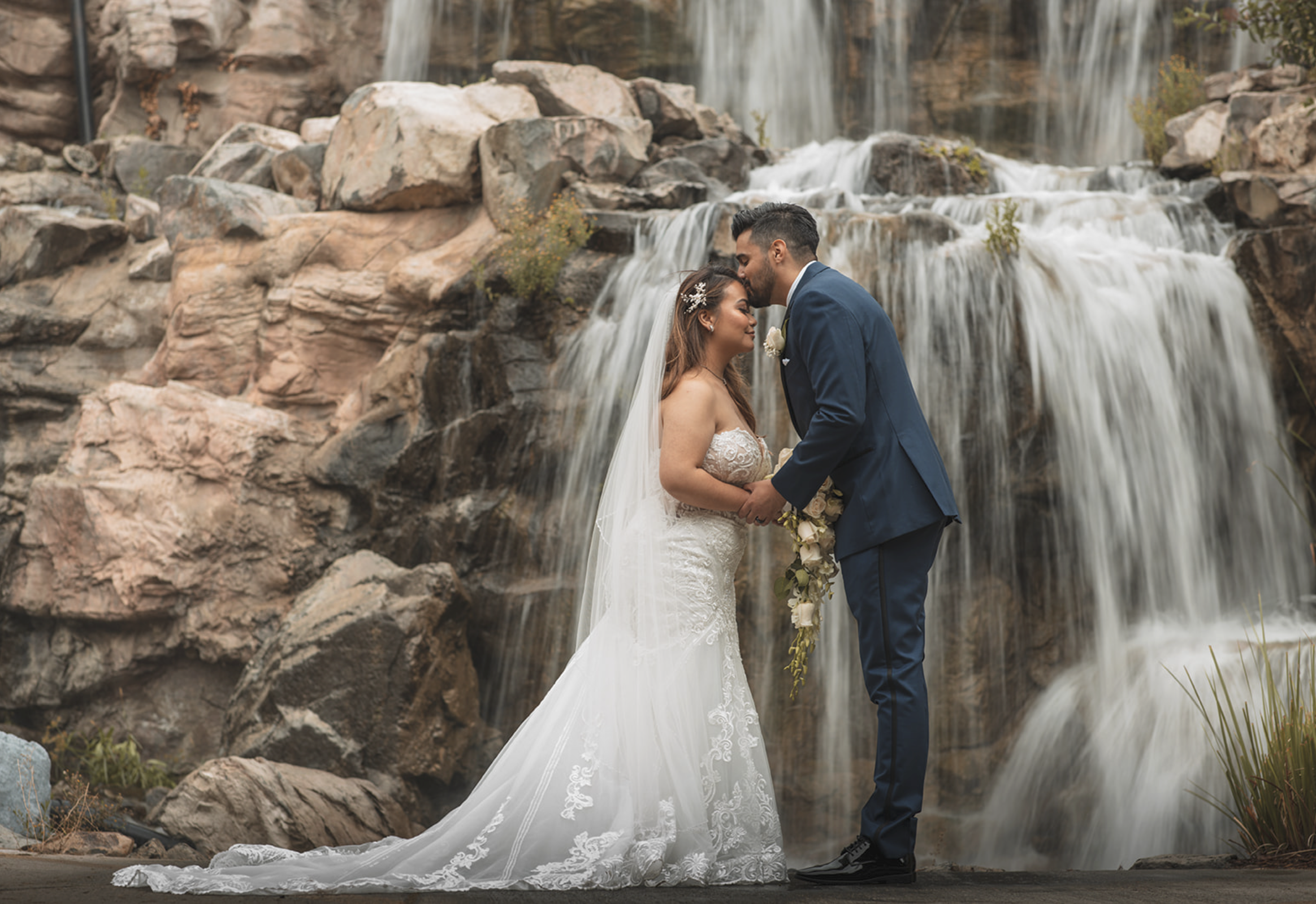 A bride and groom stand close and touch foreheads in front of a waterfall, holding hands; the bride wears a white lace wedding gown and veil, the groom wears a blue suit, and the bride holds a bouquet of white roses.