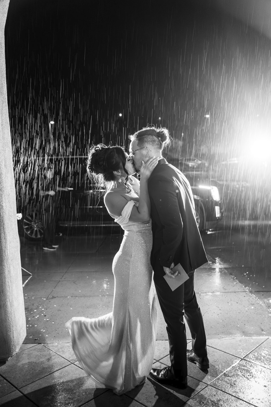 A black and white photo of a couple kissing in the rain at night, with a bright light behind them illuminating raindrops, and parked cars visible in the background.