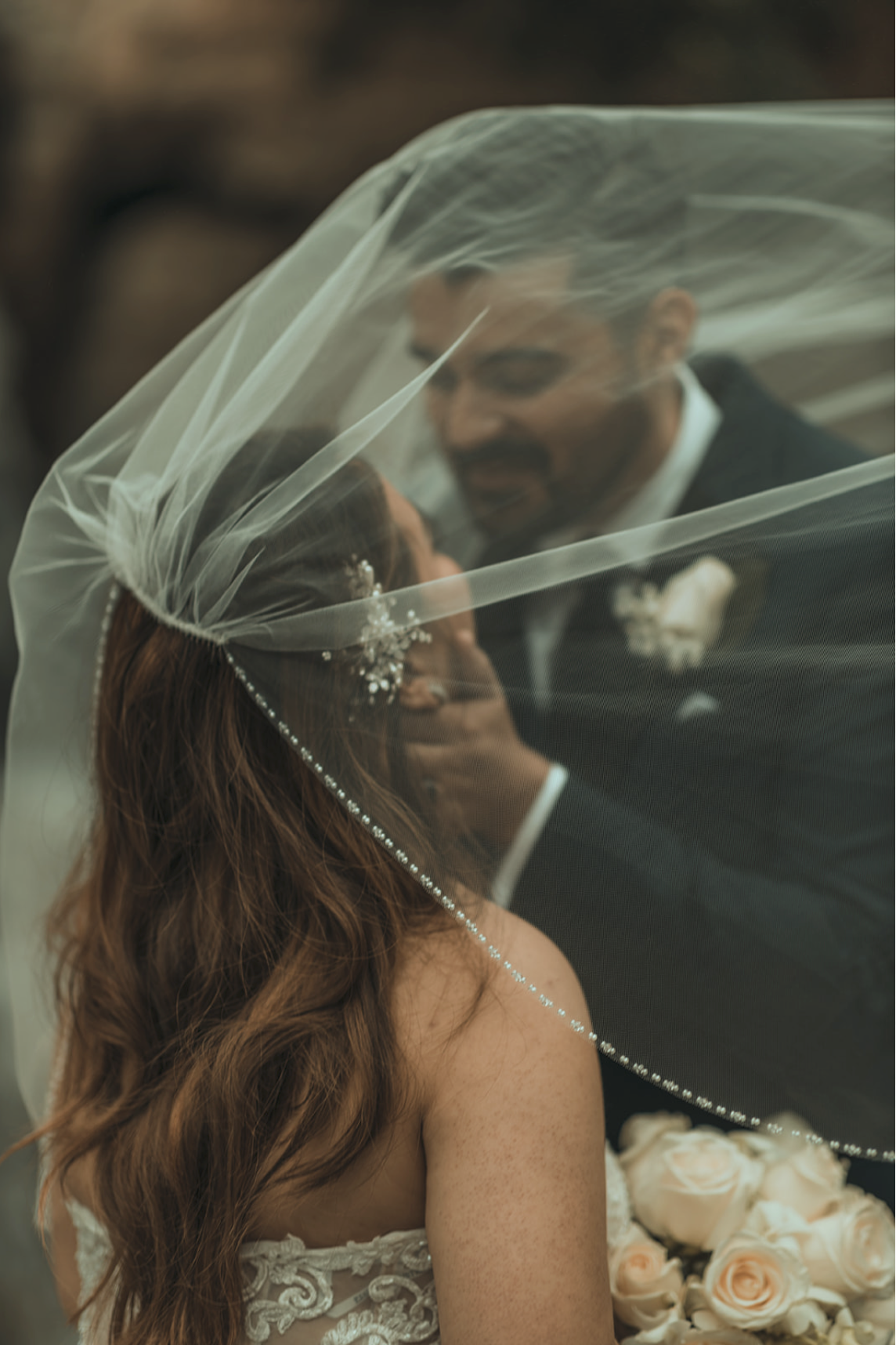 A bride and groom share a moment under a veil during their wedding ceremony, with the bride holding a bouquet of light-colored roses.