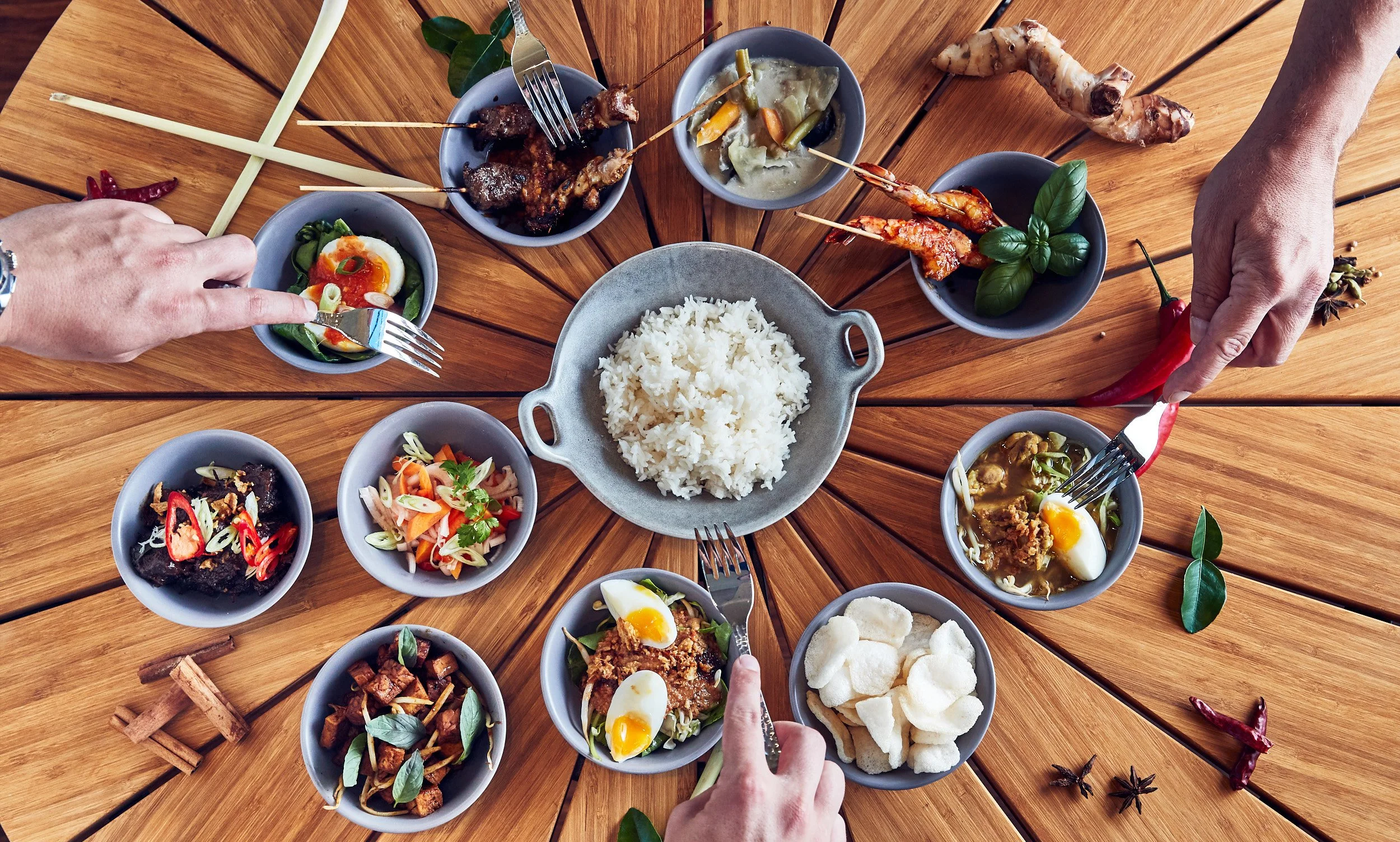A round wooden table with various Indonesian dishes in small bowls, including rice, salads, and spicy foods, with people reaching for the food using forks and chopsticks.
