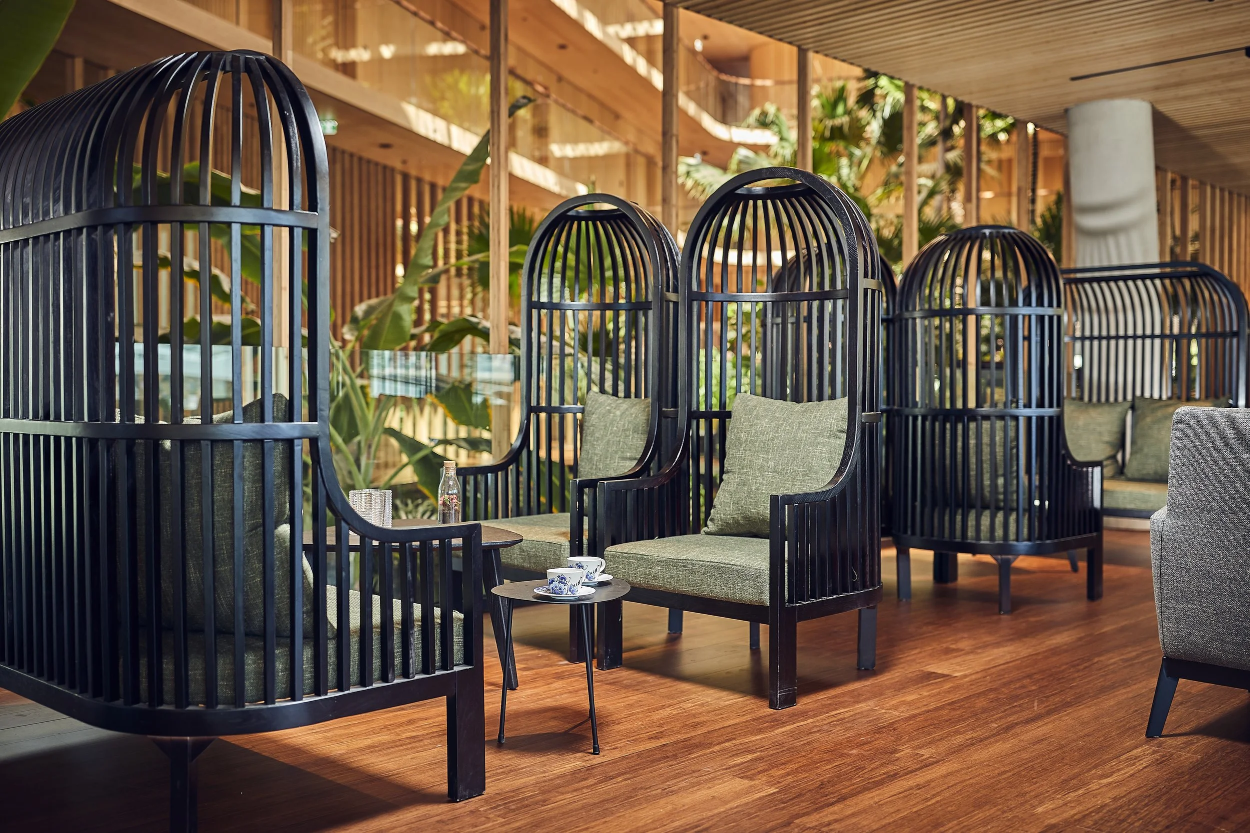 Cozy indoor lounge space with black wooden cabana chairs with green cushions, small tables with teacups and bottles, surrounded by lush green plants and wood-paneled decor.