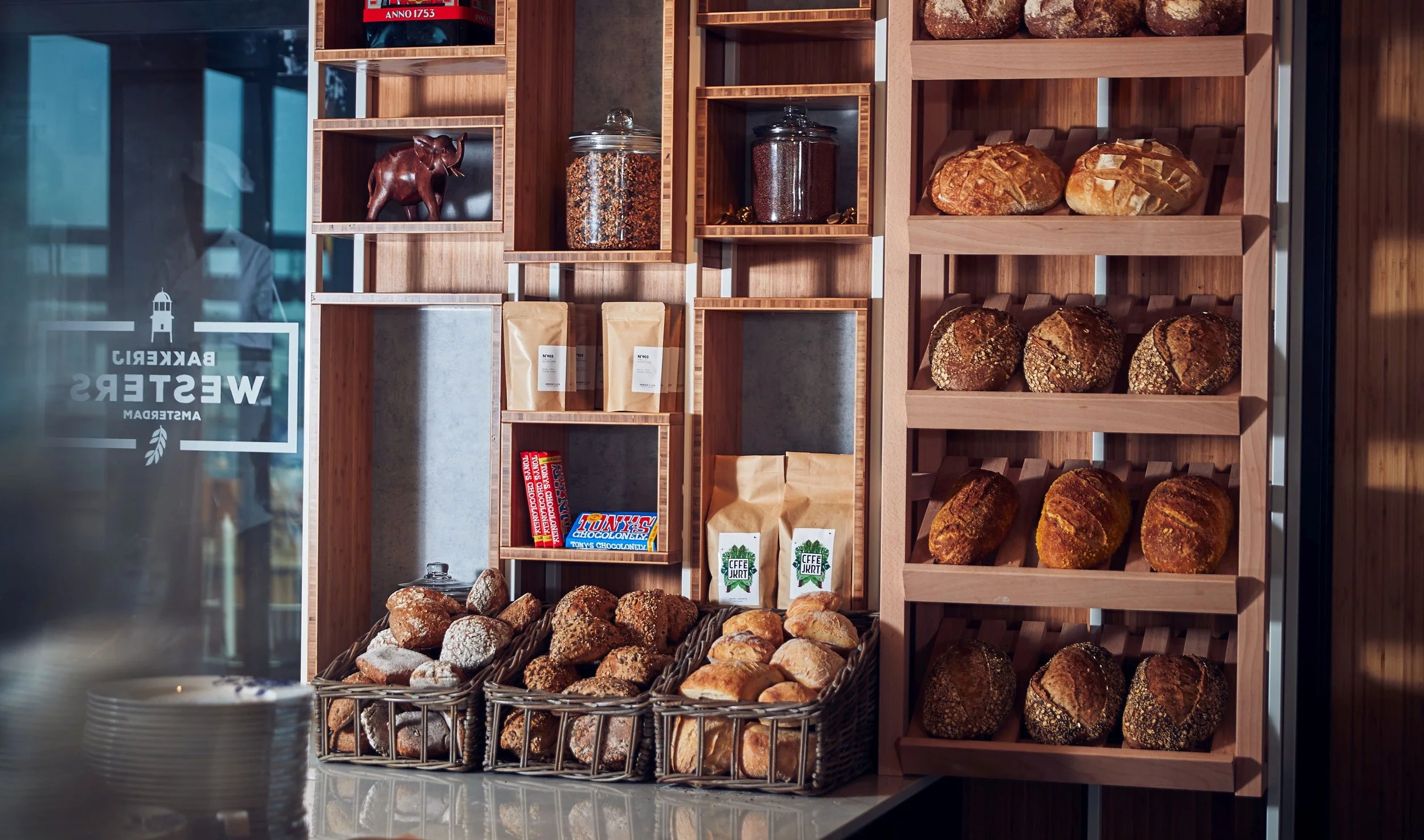 Display of assorted bread and baked goods in a bakery, with shelves holding loaves, rolls, jars, and packaged items behind a glass window.