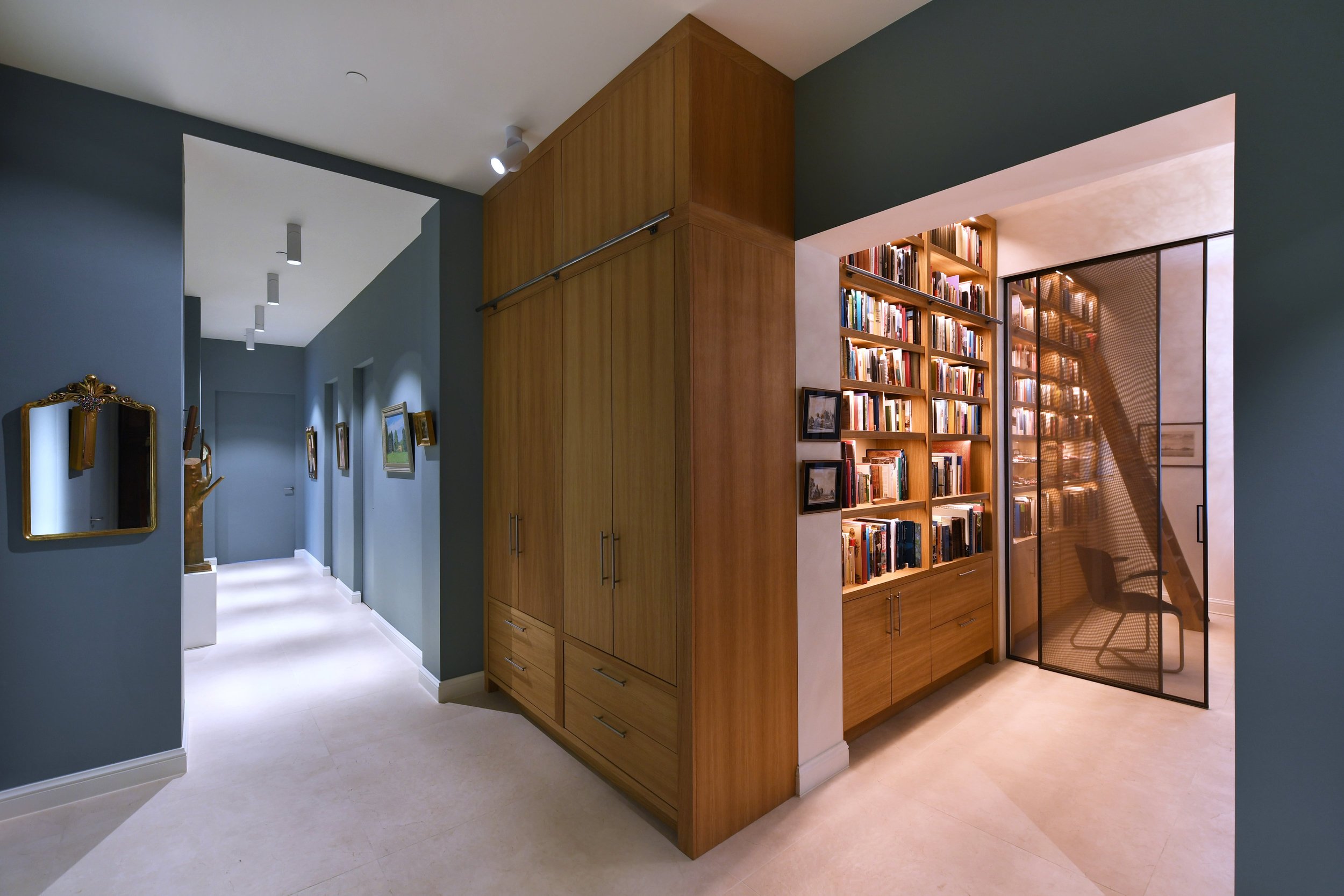 Interieur van een modern huis met blauwe muren, een gang met kunstwerken aan de muur en een boekenkast met glazen schuifdeuren, een leeshoek met een stoel en boeken.