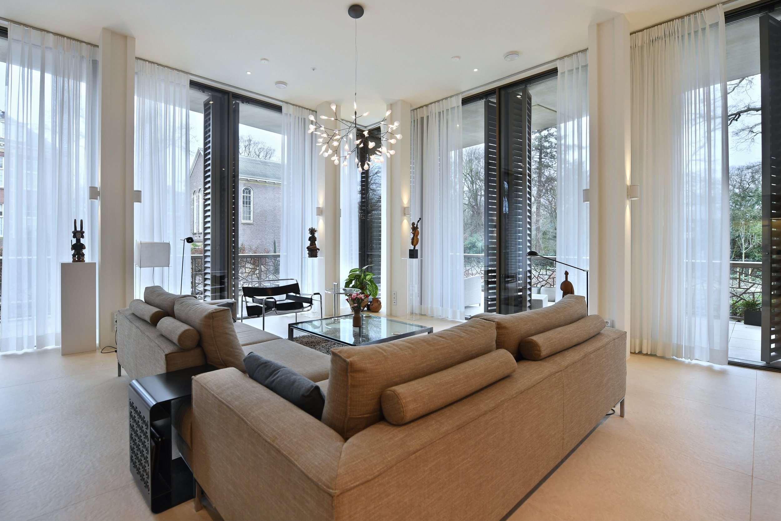 Luxe woonkamer met grote ramen, beige bank, zwarte en witte stoelen, groene plant, centrale lamp, en witte gordijnen