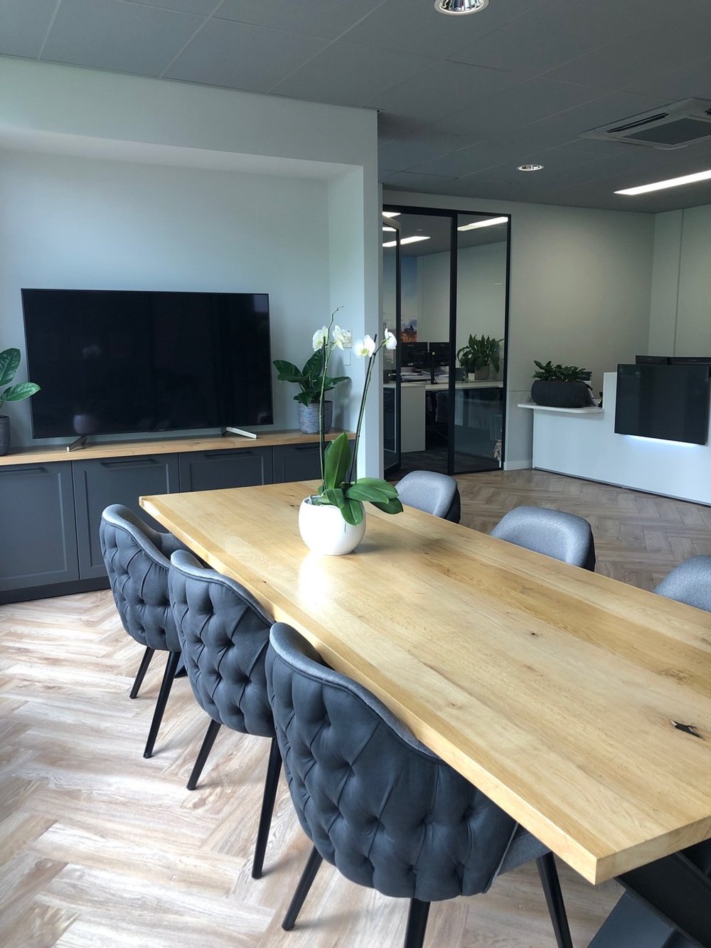 Een modern kantoorruimte met een houten vergadertafel, omgeven door grijze gestoffeerde stoelen, met een witte bloempot met orchidee op tafel, een grote tv en planten op een kast, en een gescheiden kantoorruimte met glazen deuren op de achtergrond.