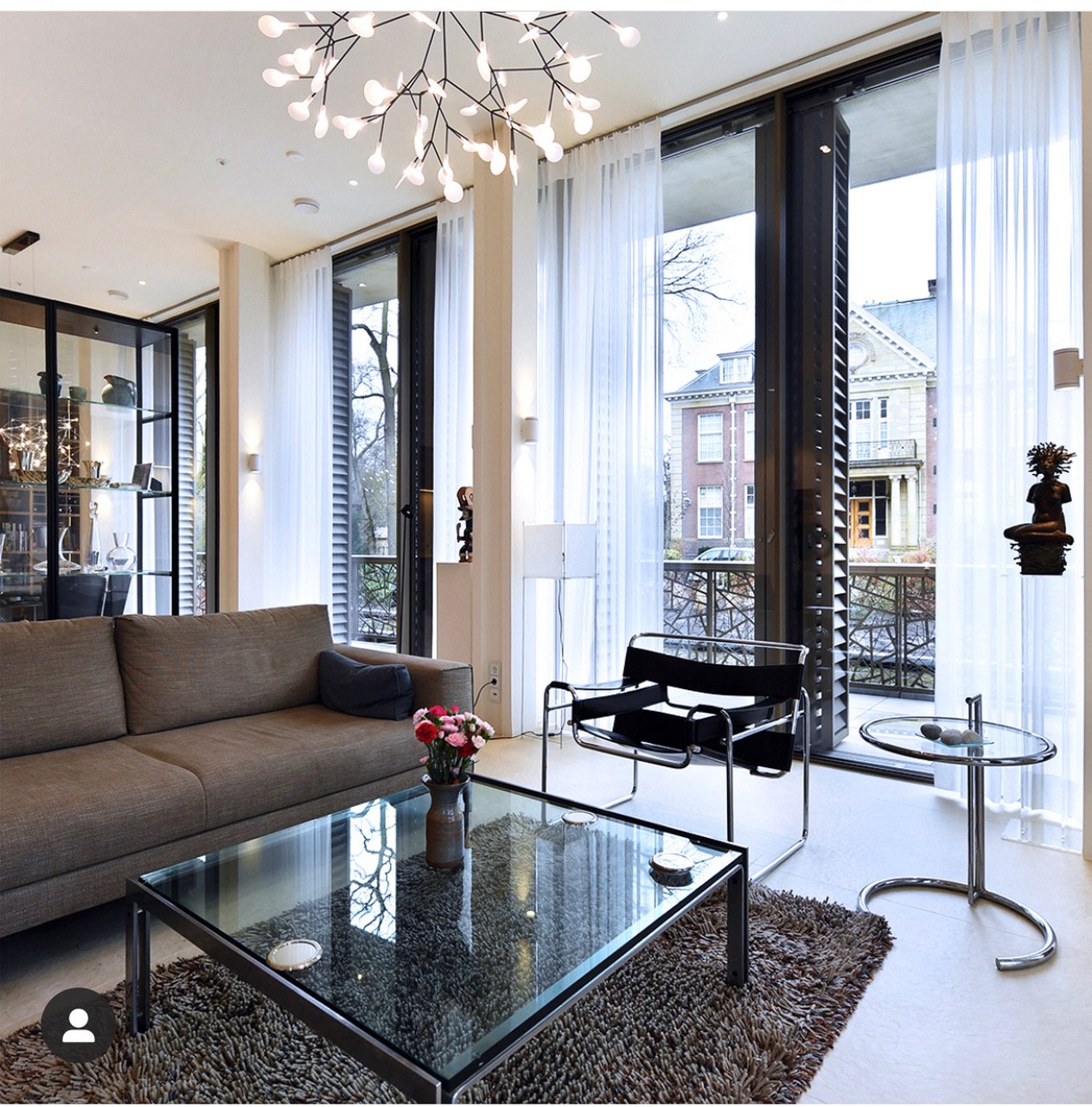 Woonkamer met grote ramen en witte gordijnen, een bruine bank, een zwarte Scandinavische stoel, een glazen salontafel met bloemen en decoraties, en een stijlvolle kroonluchter.