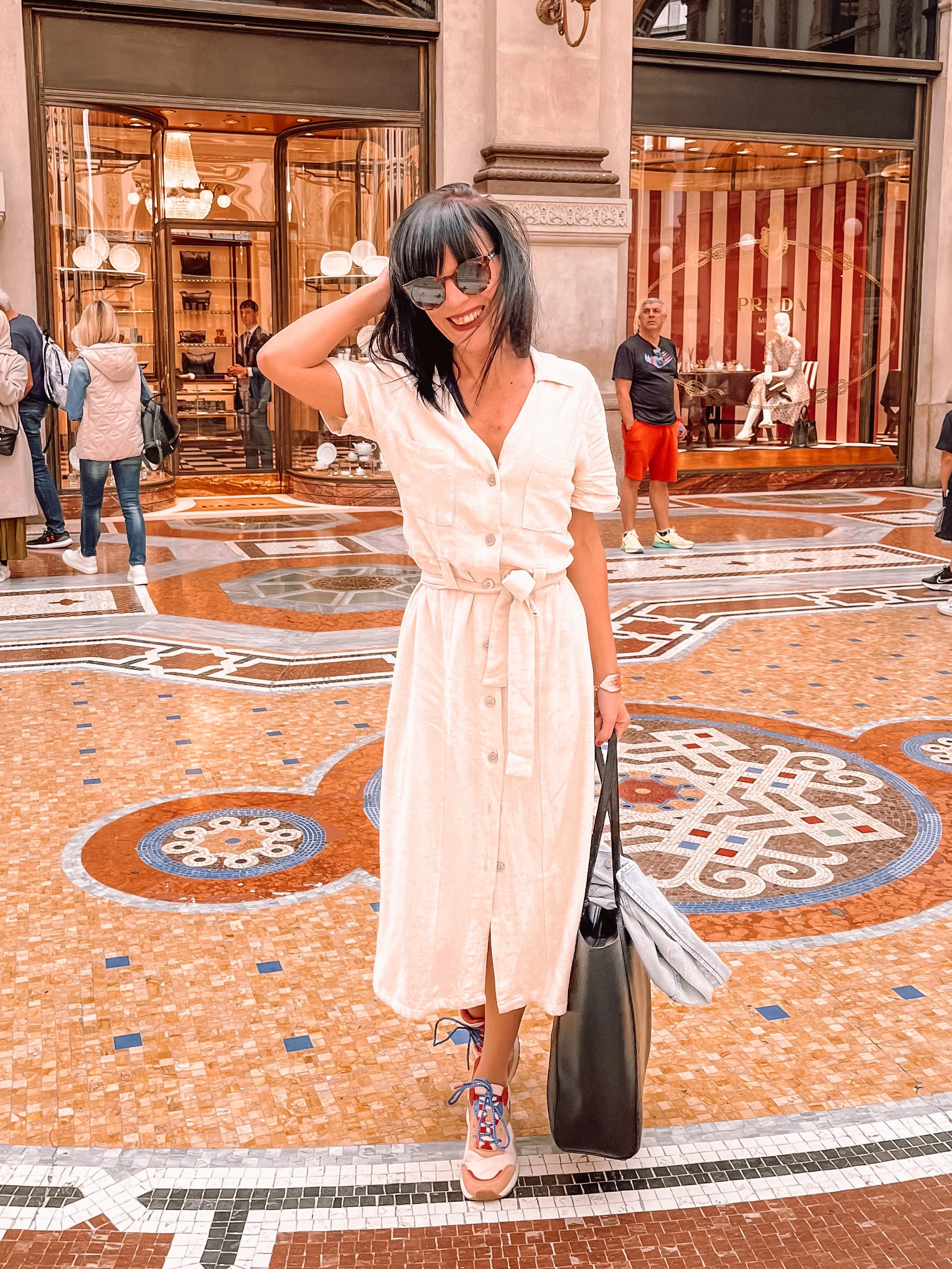 A woman with black hair wearing sunglasses and a cream-colored button-up dress with a belt, standing inside a shopping galleria, holding a large black shopping bag, and smiling.