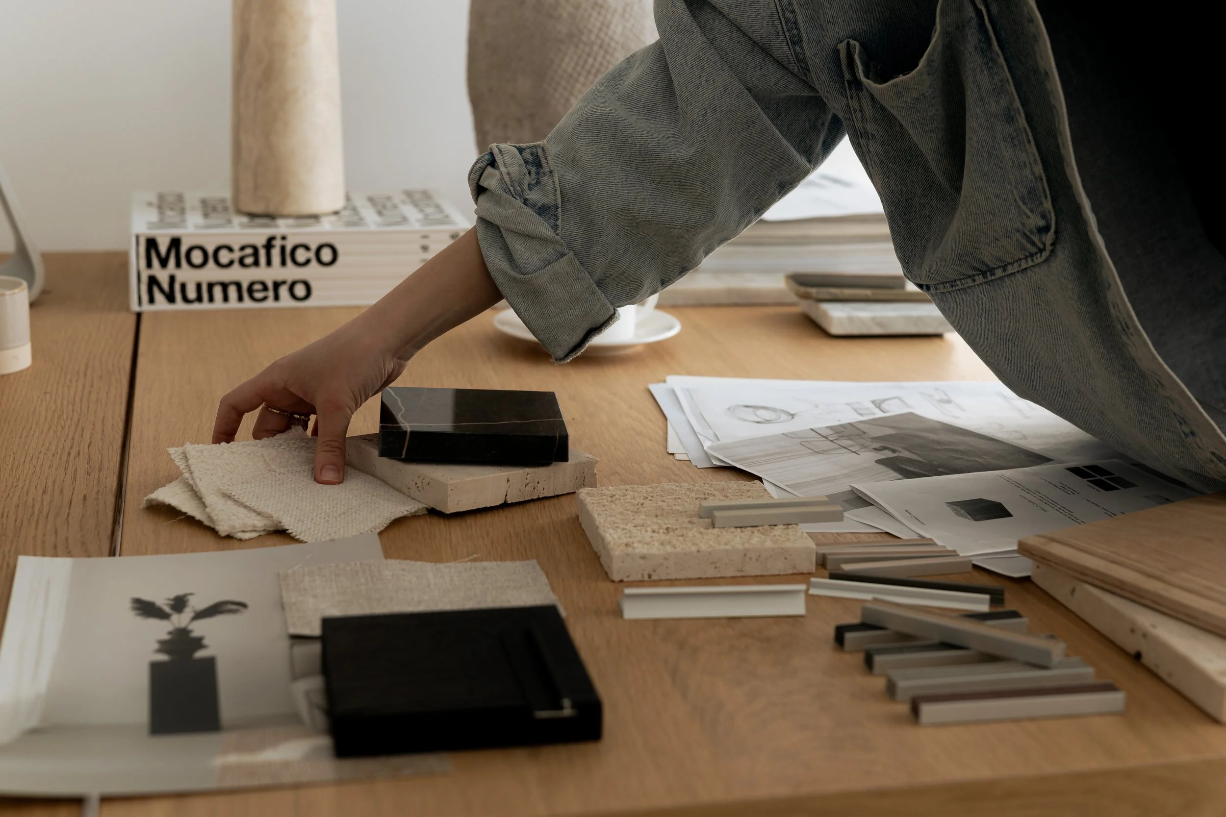 Person arranging color and material samples on a wooden table, surrounded by design plans and magazines in a workspace.