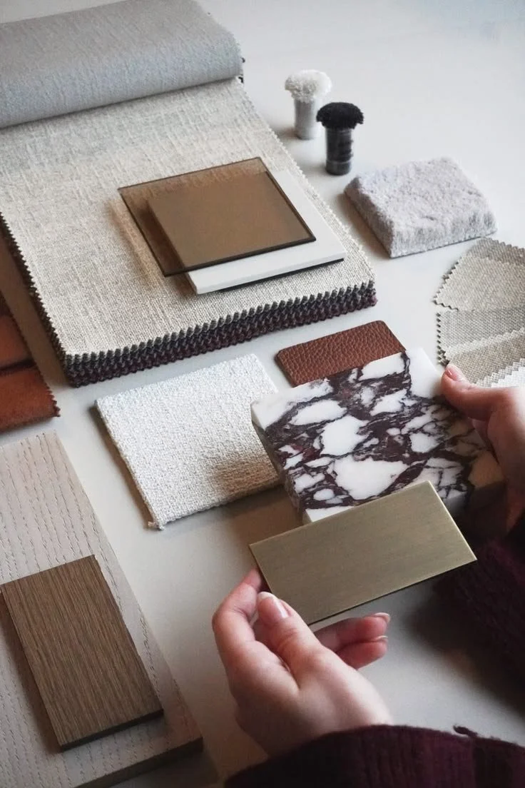 A person holds a small marble-patterned tile and a gold-colored tile sample, with various fabric swatches, color samples, and tile samples spread on a table for interior design planning.