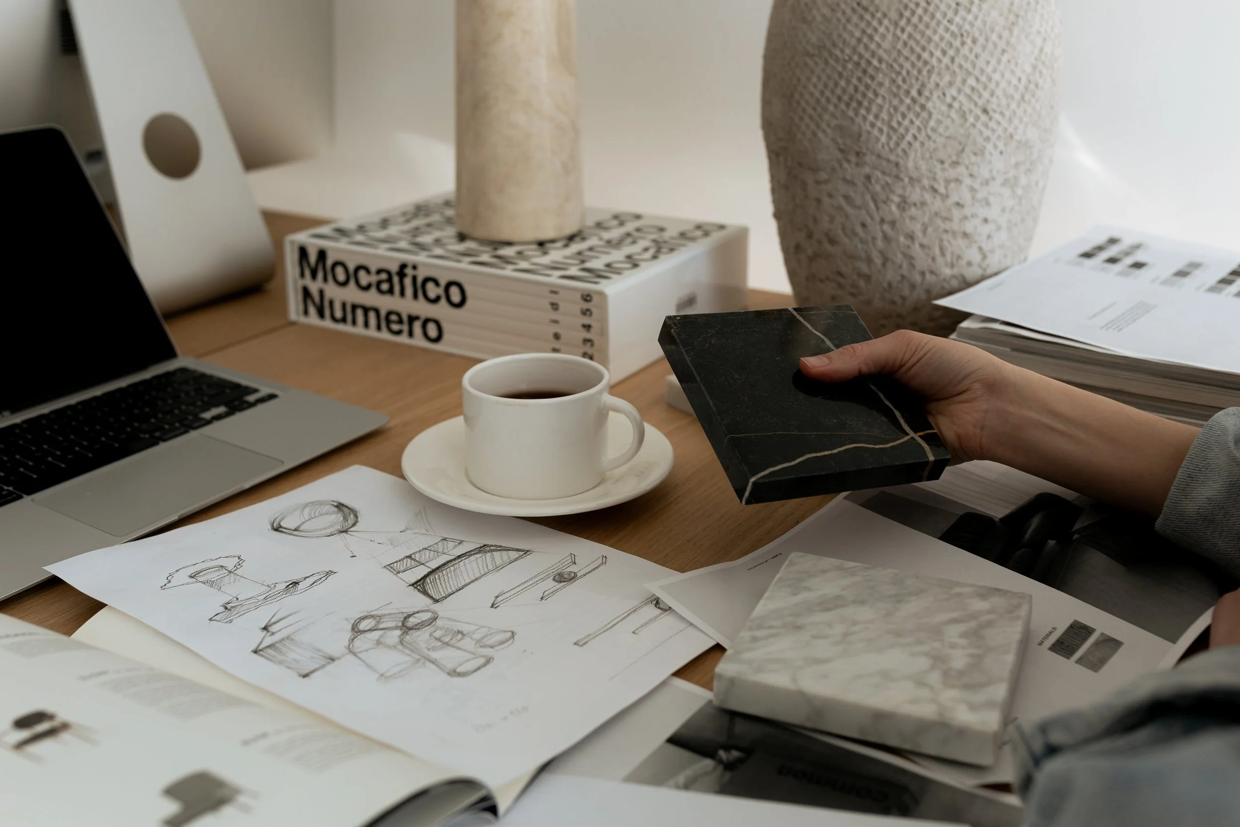 A cluttered workspace with a laptop, sketches, a cup of coffee, and color sample books on a wooden desk.