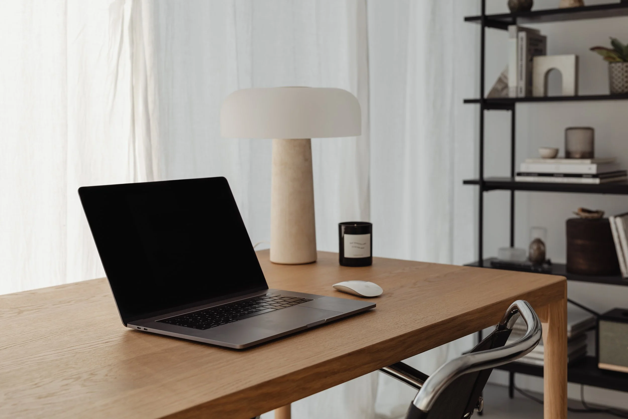 Modern home office with a wooden desk, open laptop, wireless mouse, black candle, table lamp, and a black shelving unit with decor items.