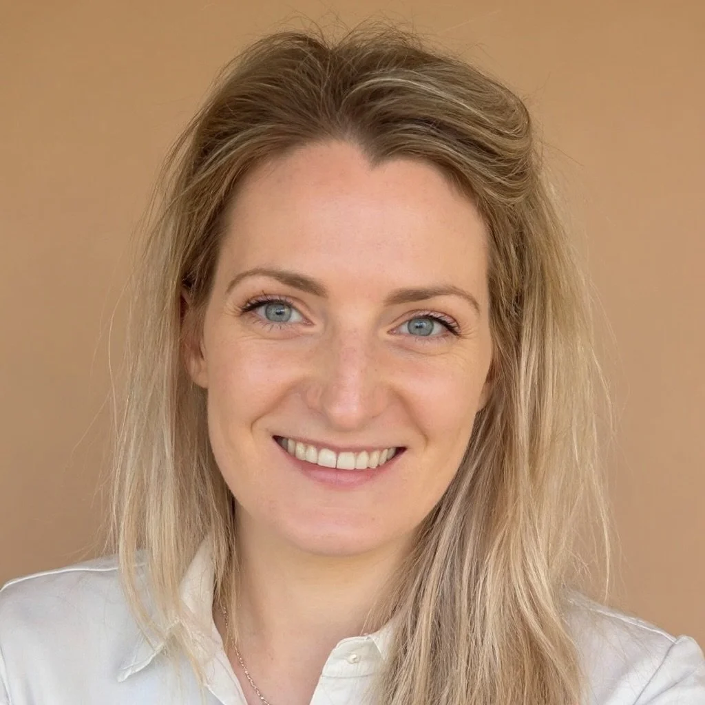 A smiling woman with long, wavy blonde hair and blue eyes, wearing a white shirt, standing against a tan background.