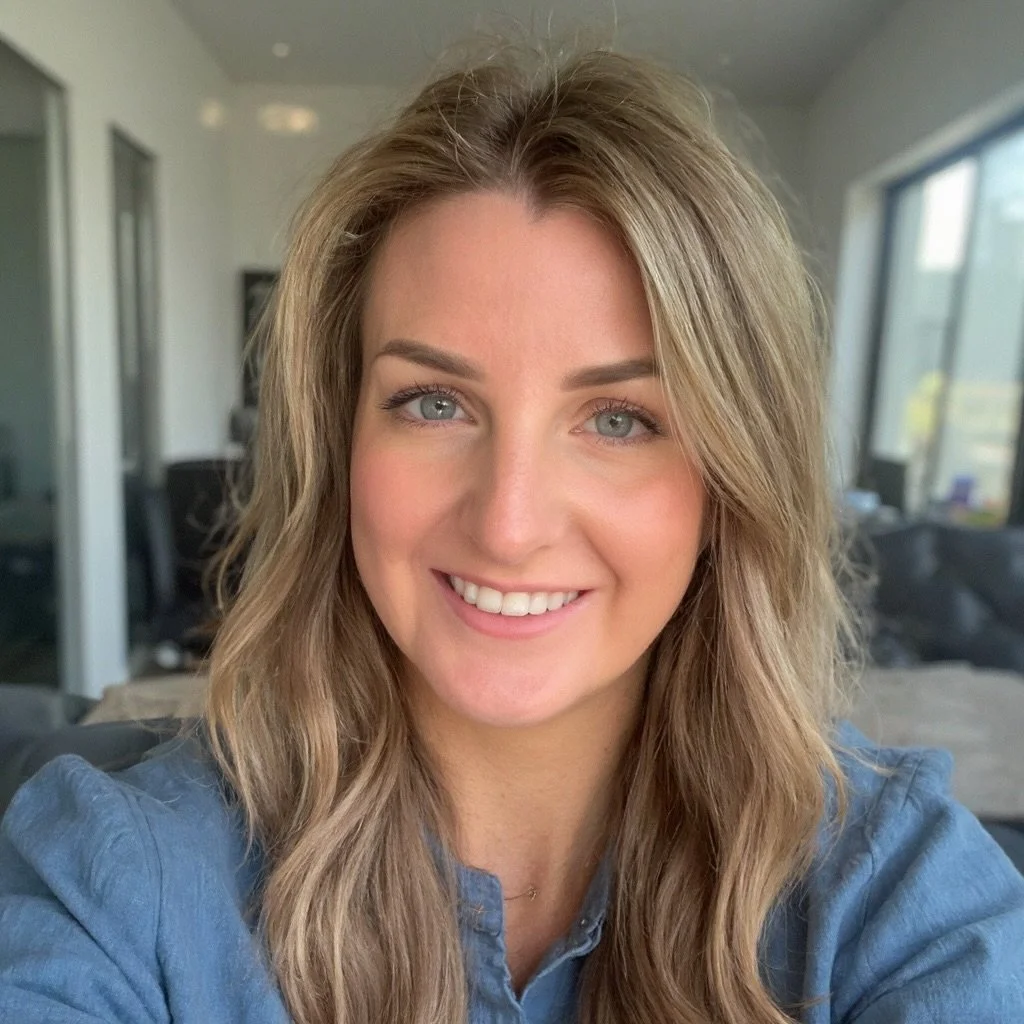 A woman with long, wavy, light brown hair and blue eyes smiling at the camera, indoors with large windows in the background.