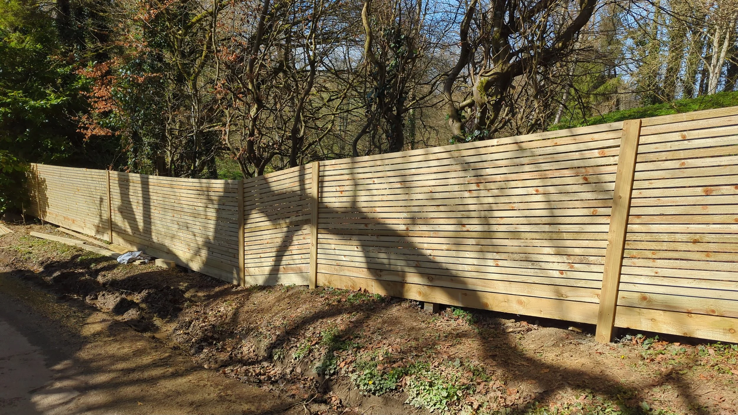 New wooden fence built along a dirt ground with trees casting shadows in the background.