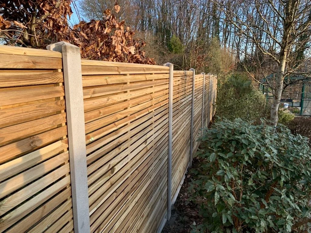 A newly installed wooden privacy fence along a garden area with green bushes and trees, under a clear sky.