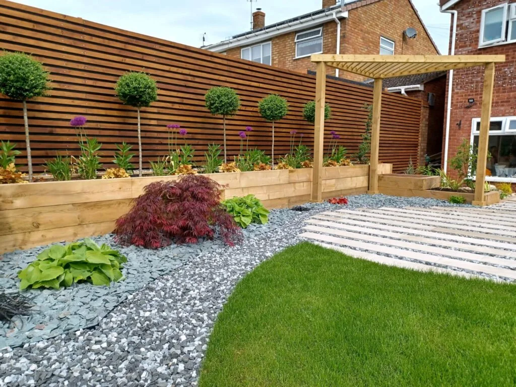 Well-maintained backyard with a lawn, gravel pathway, wooden raised bed with plants, and wooden pergola, surrounded by a horizontal wooden fence.