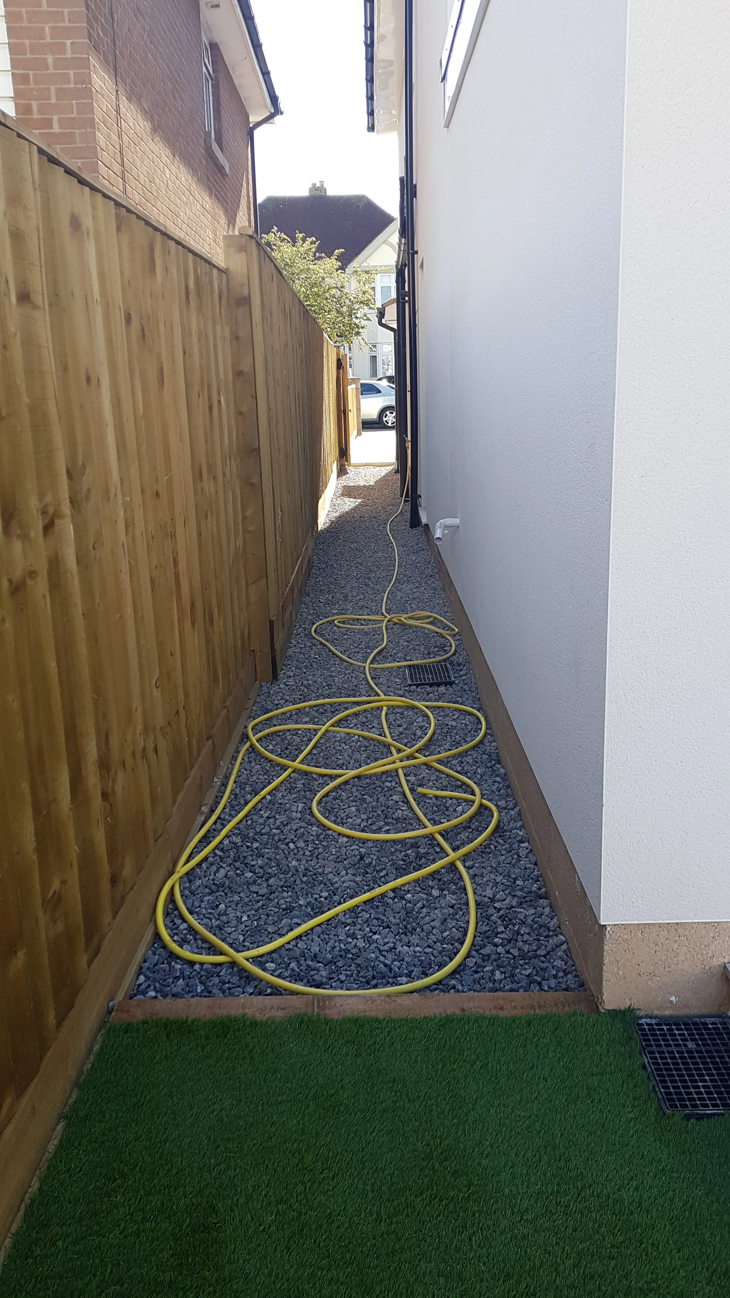 A narrow outdoor walkway between a white house and a wooden fence, covered with gravel, with yellow extension cords lying on it and a patch of artificial grass at the foreground.