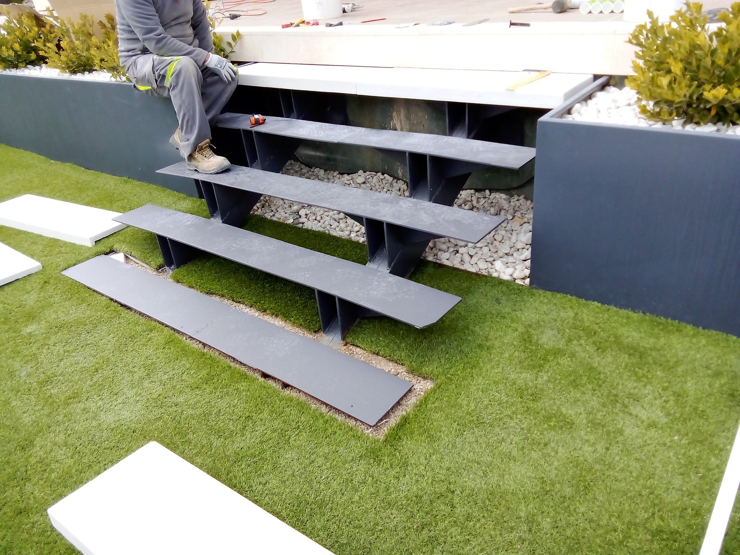 Persona trabajando en la instalación de escalones de metal negro en un jardín con césped y plantas decorativas.