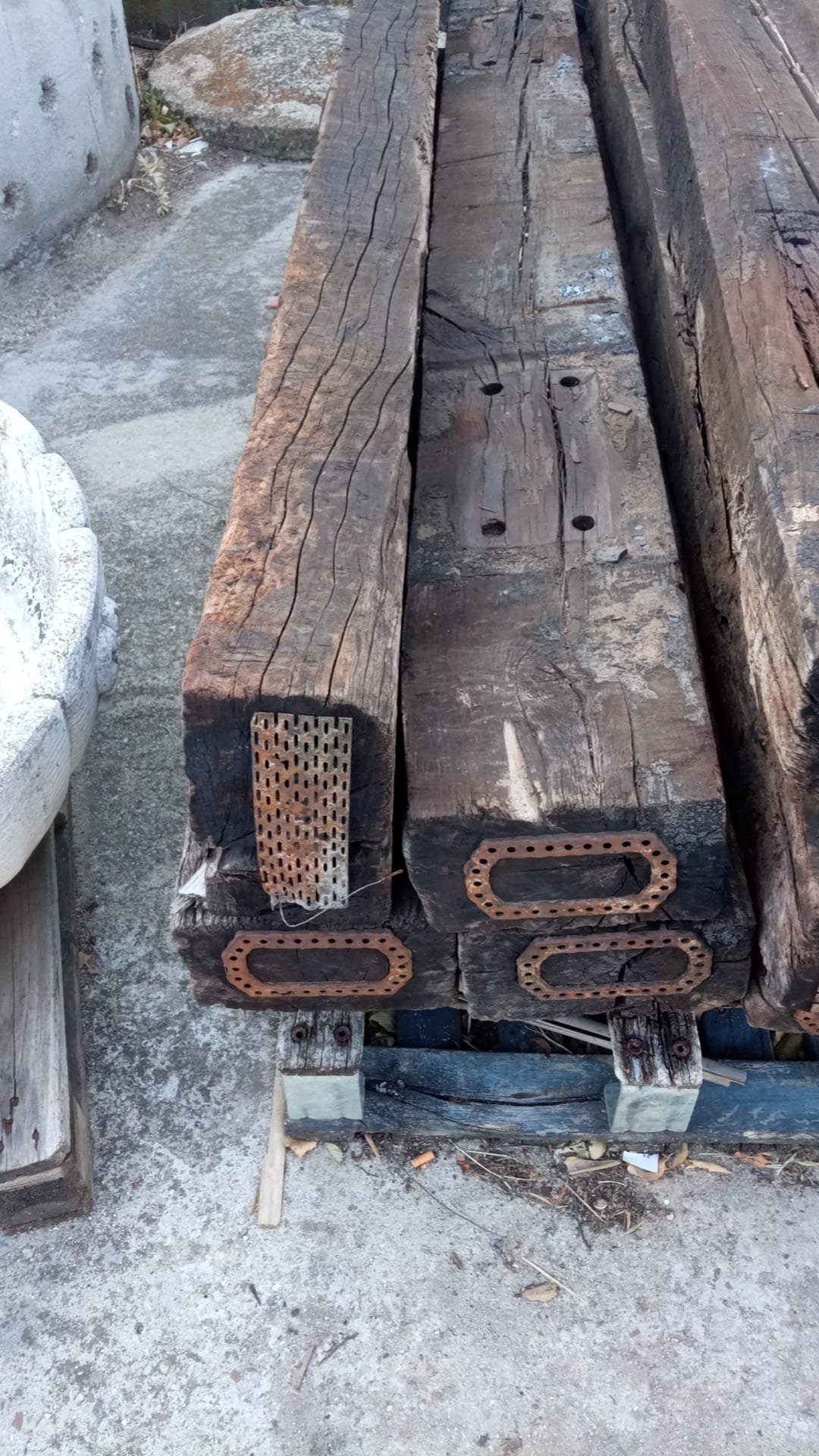 Listones de madera con molduras de hierro en sus extremos, en un entorno de suelo de concreto y elementos de construcción alrededor.