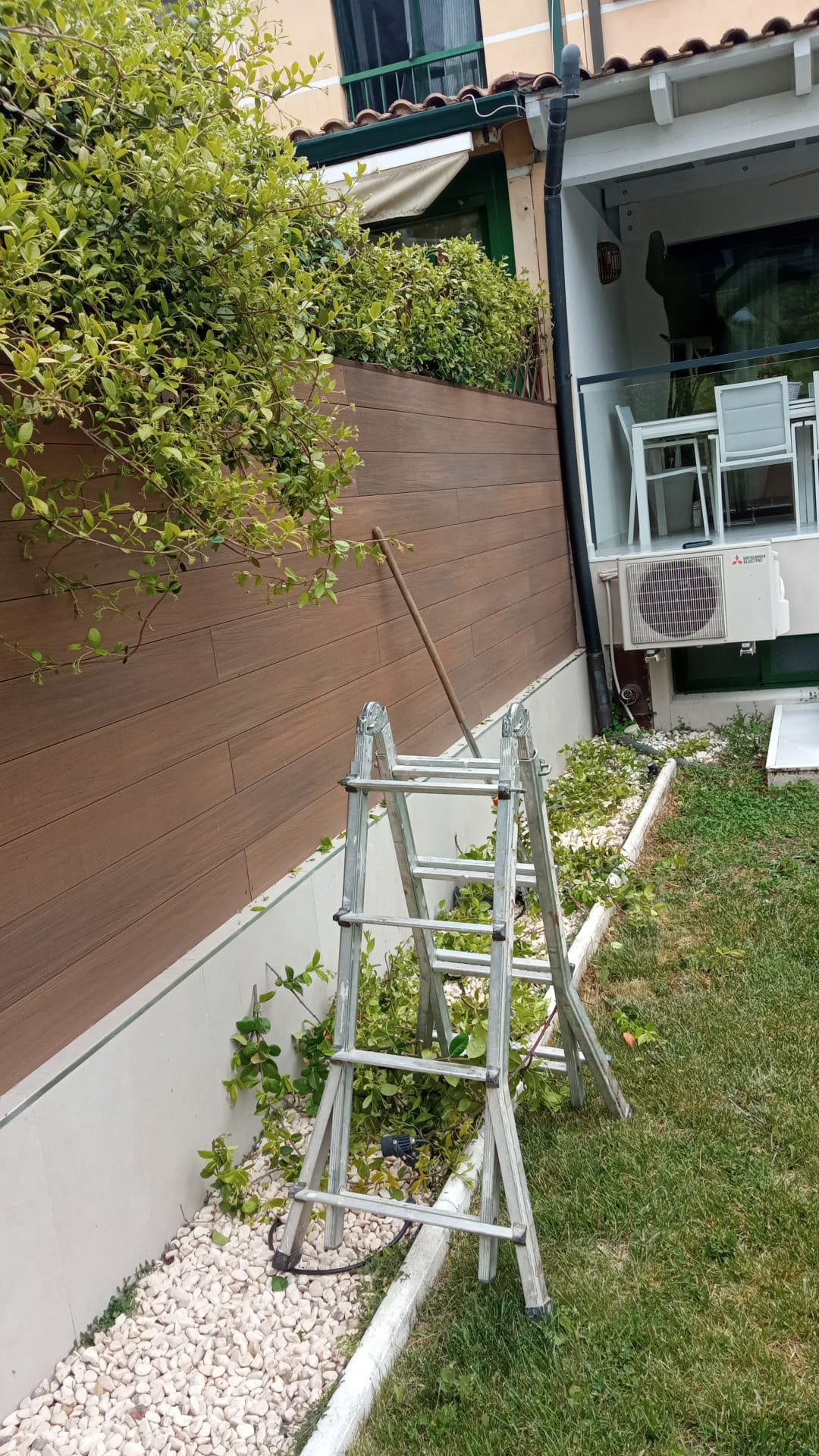 Una escalera de aluminio descansando sobre piedras blancas en un jardín con plantas y césped, junto a una cerca de madera y una pared de una casa.