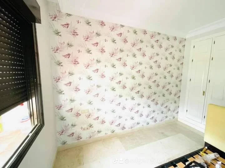 Habitación con pared decorada con papel tapiz de flores y plantas en tonos rosas y verdes, armario empotrado blanco y una ventana con persiana negra.