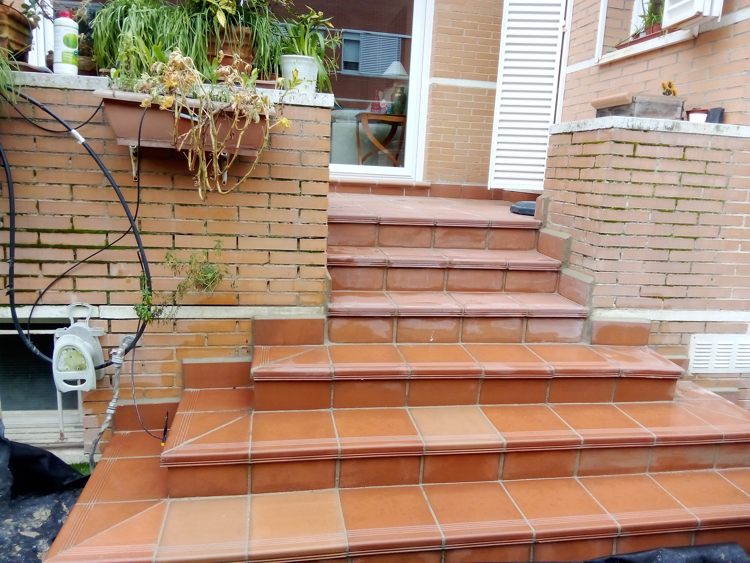 Escalones de terraza con baldosas de color terracota y paredes de ladrillo, con plantas en la parte superior y una puerta de vidrio con marco blanco al fondo.