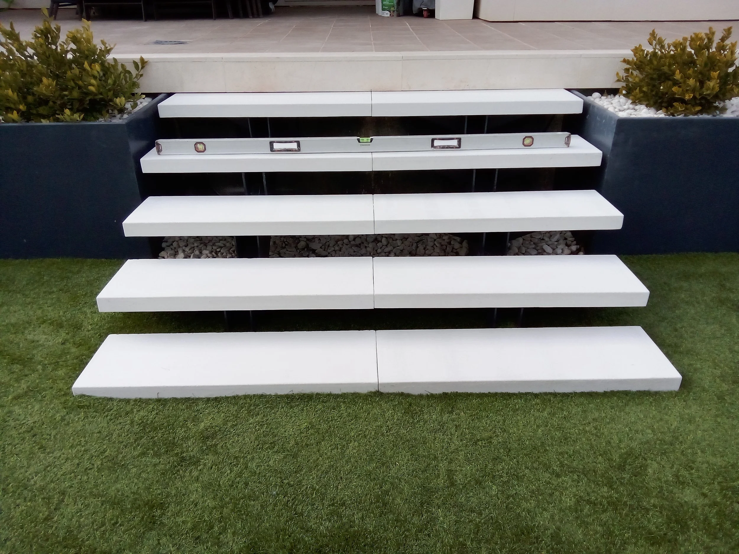 Escalera moderna de tres peldaños blancos en un jardín con césped, con niveladores y un fondo de planta con piedras decorativas.