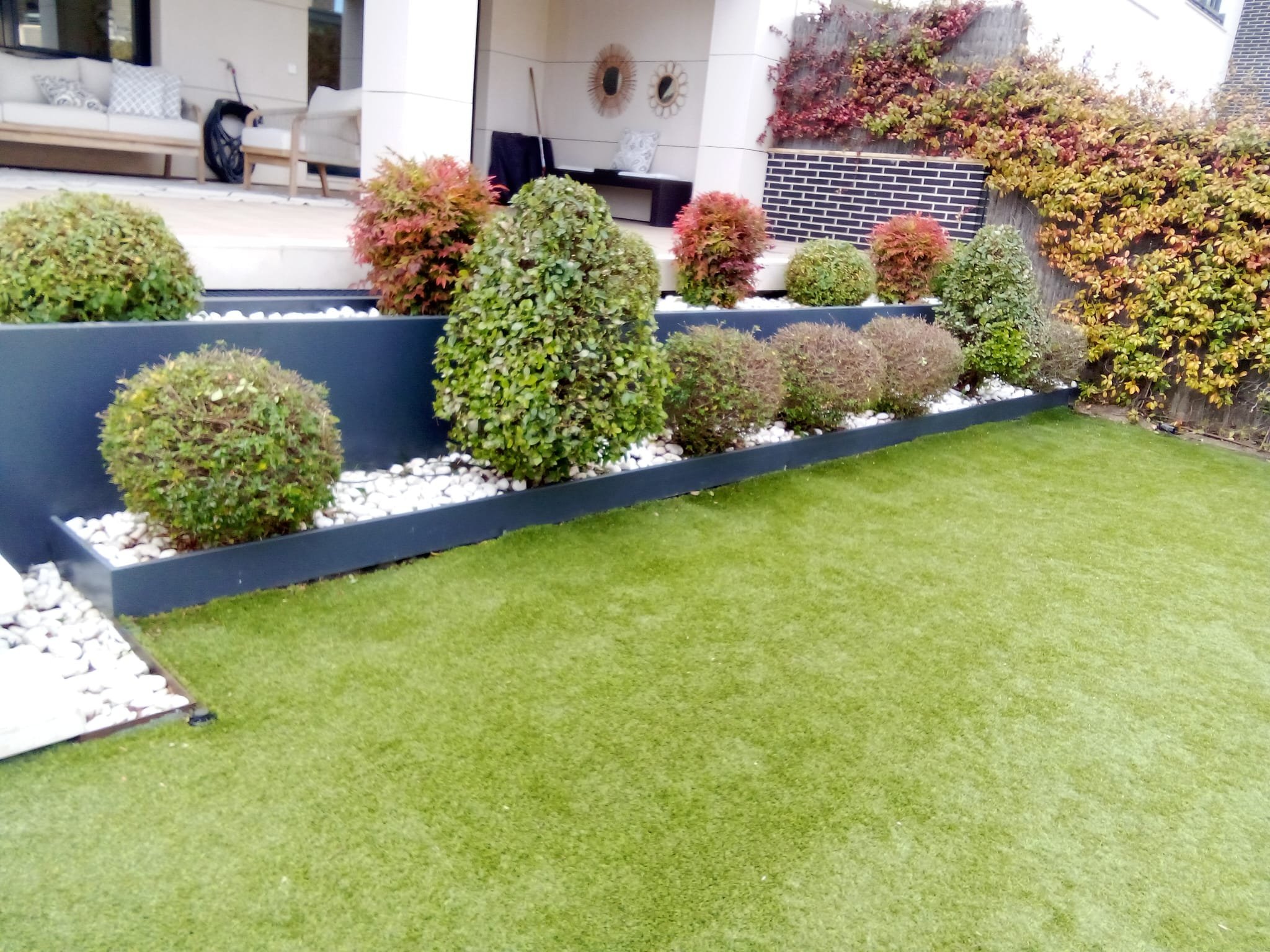 Jardín con césped artificial, macizos de arbustos y plantas con colores otoñales, zona de terraza y muebles en el fondo.