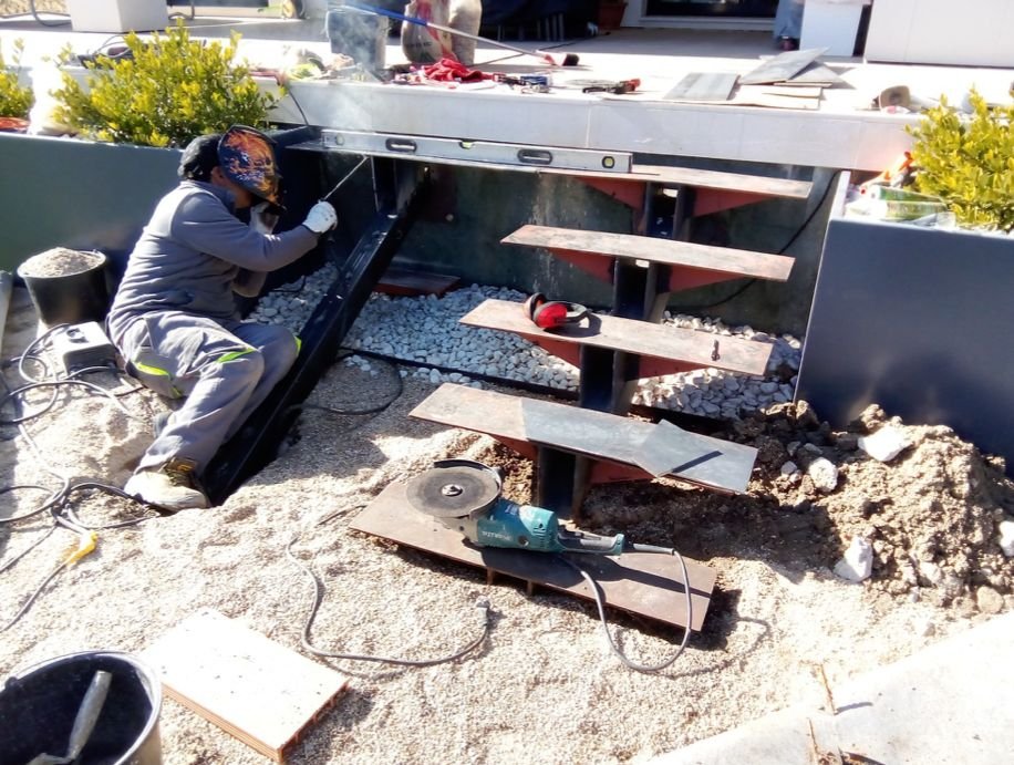 Un trabajador en una obra de construcción, soldando un marco metálico junto a unas escaleras metálicas en un espacio exterior, con herramientas y materiales de construcción alrededor.