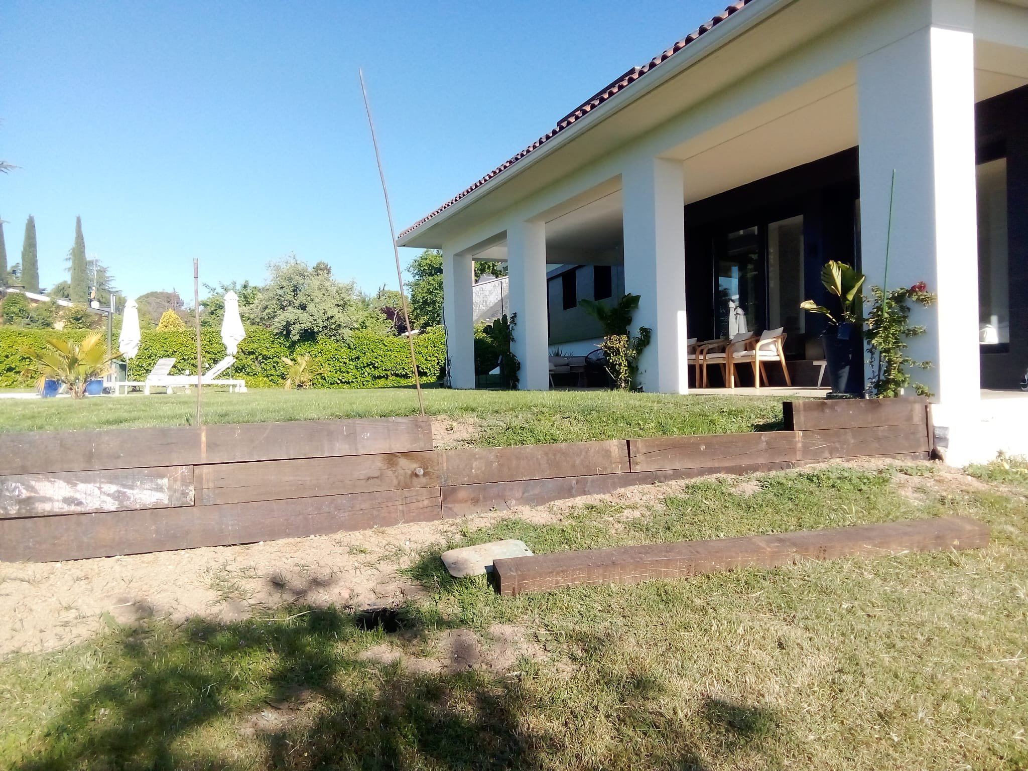 Vista de un patio de casa con césped, muebles de exterior y plantas, con una casa moderna de fondo.