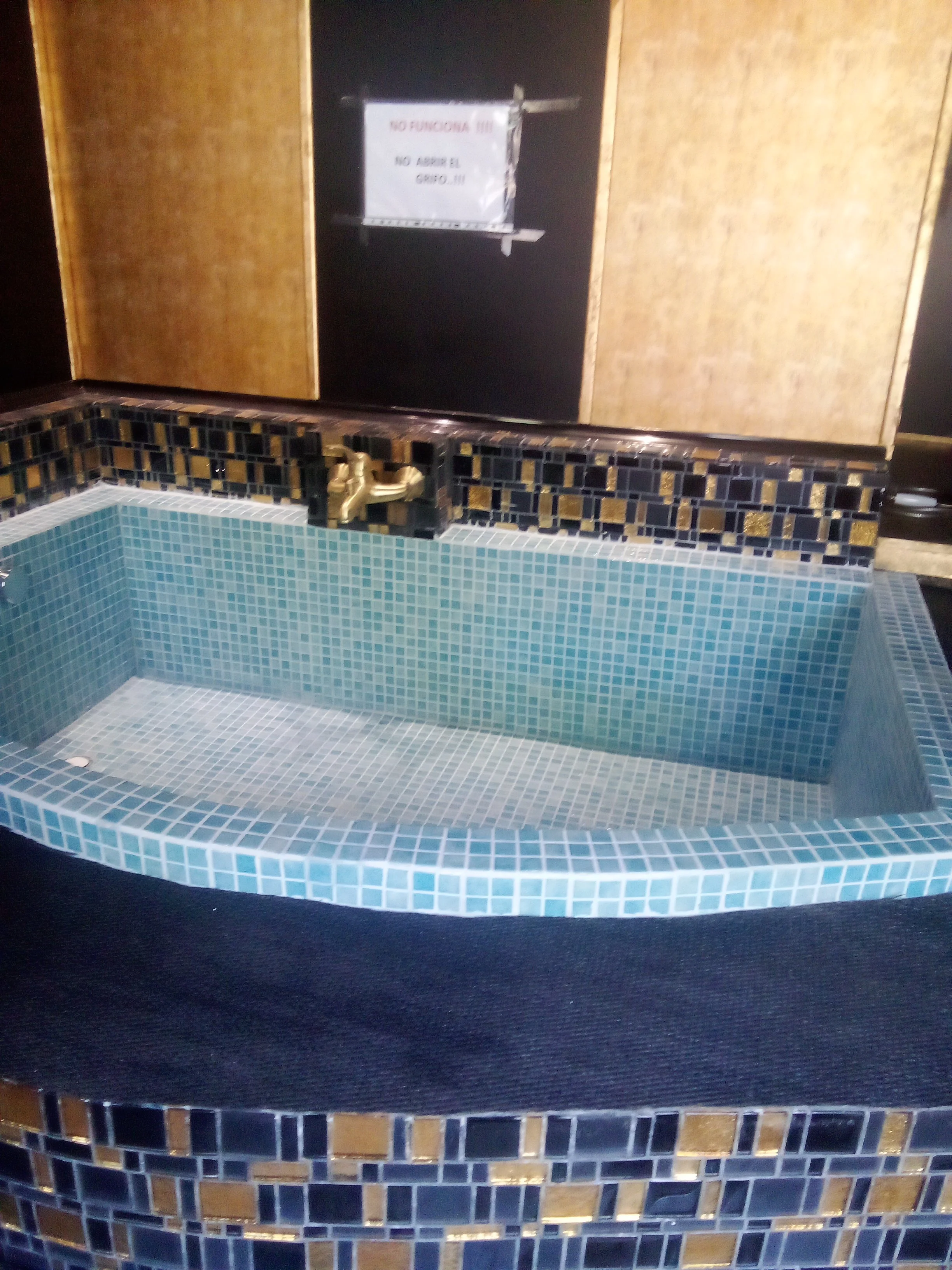 Bañera de azulejos en tonos azules y dorados con grifo dorado, y cartel que dice "No funciona, no abrir el grupo".