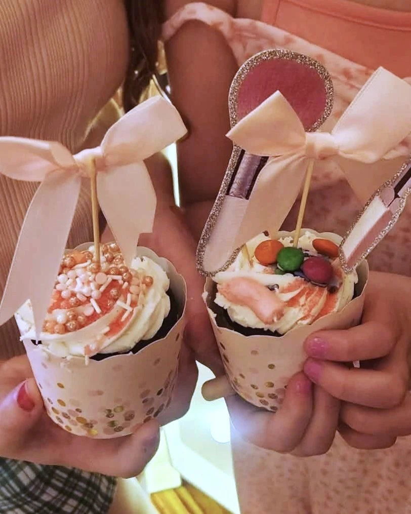 Pink and white cupcakes with decorative toppers, displayed on a three-tiered acrylic stand with pink lighting, against a wooden background.