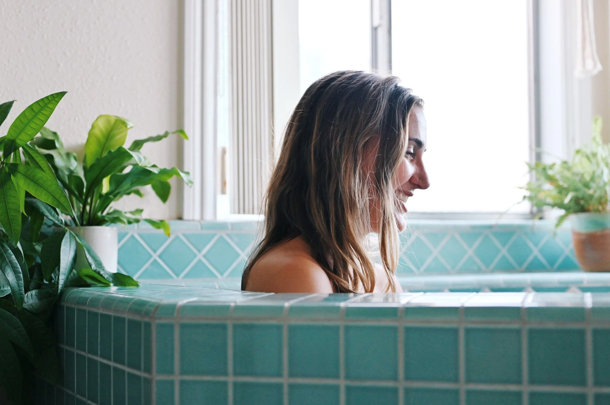 A woman enjoying a relaxing bath in a tiled bathtub with natural light coming through the window, surrounded by houseplants.