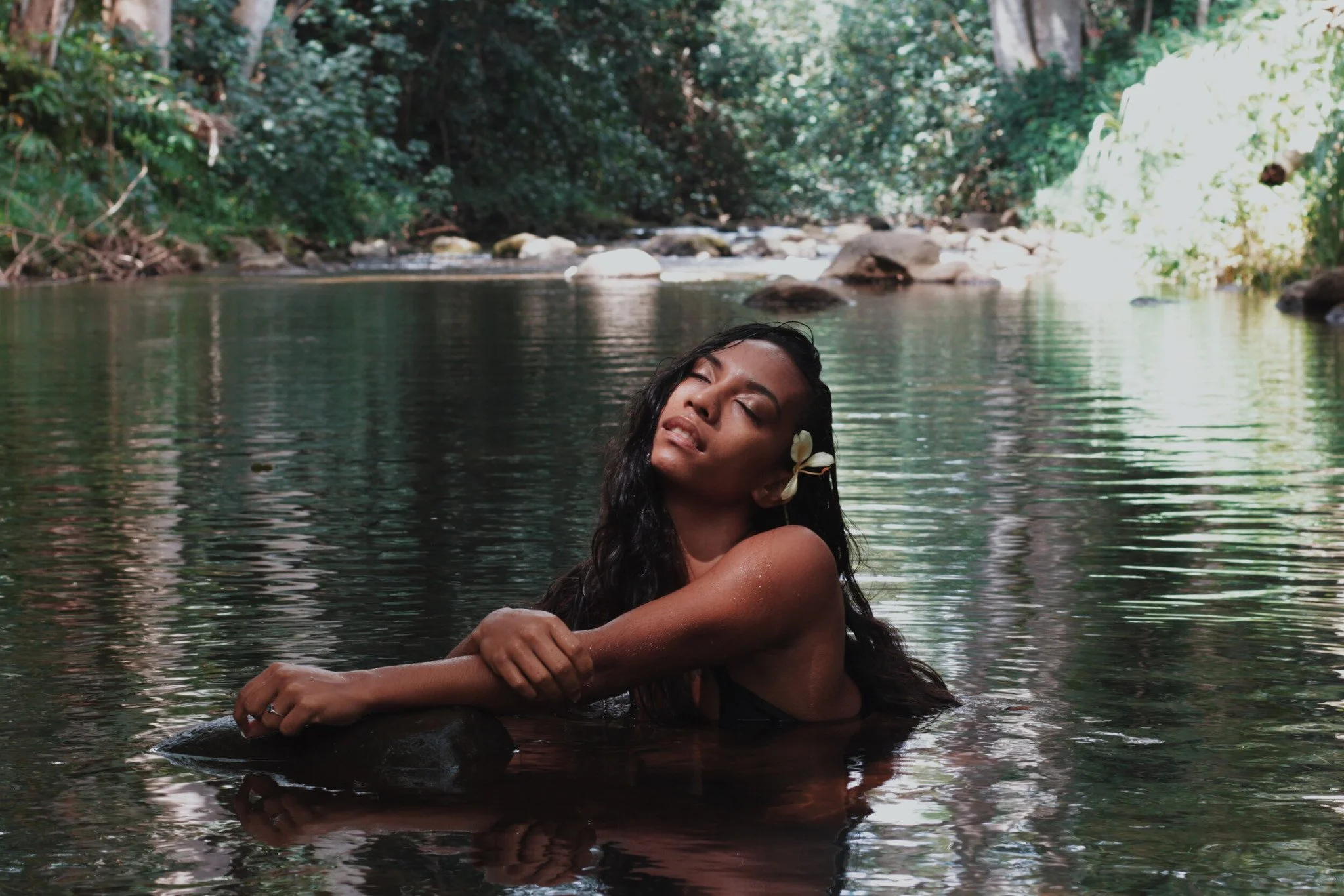 A woman with a flower in her hair relaxes in a river surrounded by lush greenery.