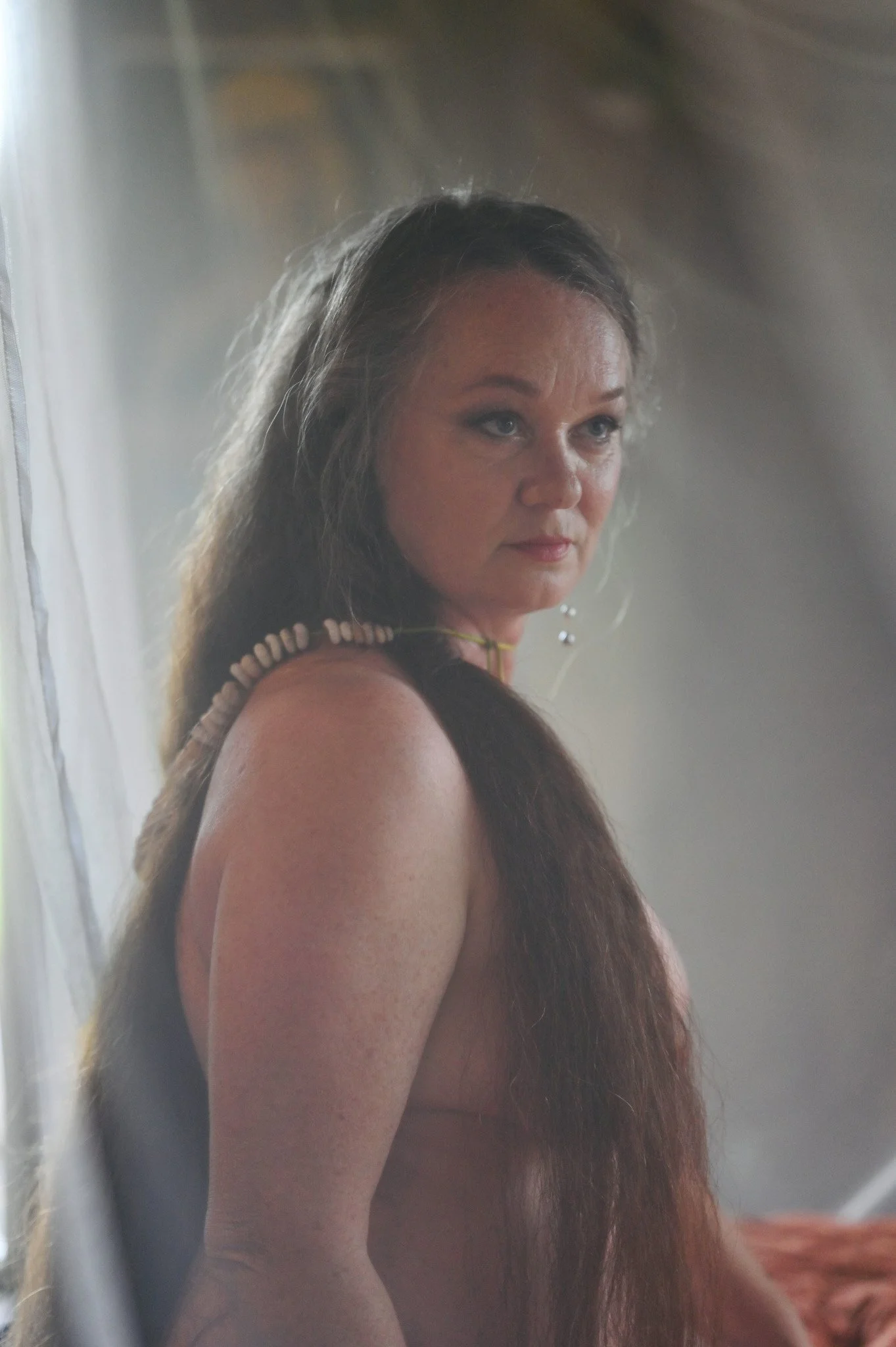 A woman with long dark hair and light skin, wearing a shell necklace and earrings, looking over her shoulder with a neutral expression.