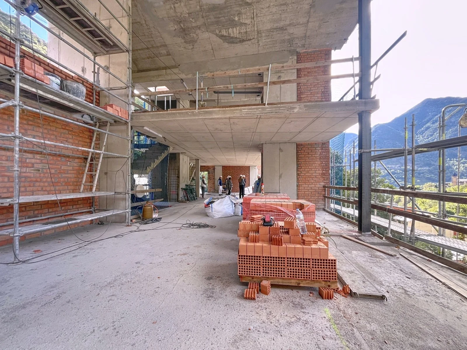 Construcción en progreso en un edificio, con estructuras de metal y ladrillos apilados, y un grupo de trabajadores en el fondo. Las paredes todavía están en construcción y hay materiales y herramientas esparcidos en el sitio.