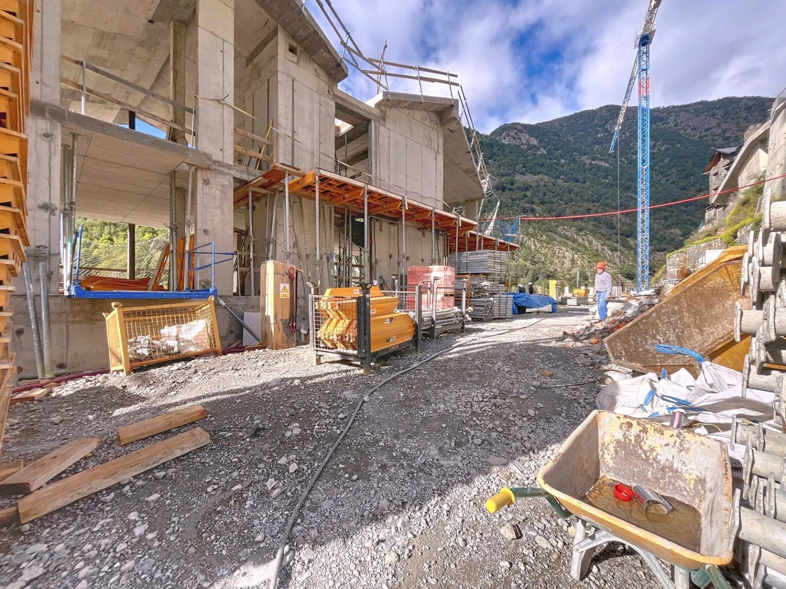 Sitio de construcción con estructura de hormigón en construcción, maquinaria y materiales de construcción, cielo parcialmente nublado y montañas en el fondo.