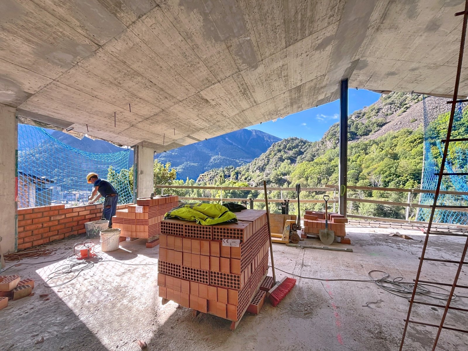 Construcción de un edificio con vista a montañas y árboles, con bloques de ladrillos y herramientas en el sitio.