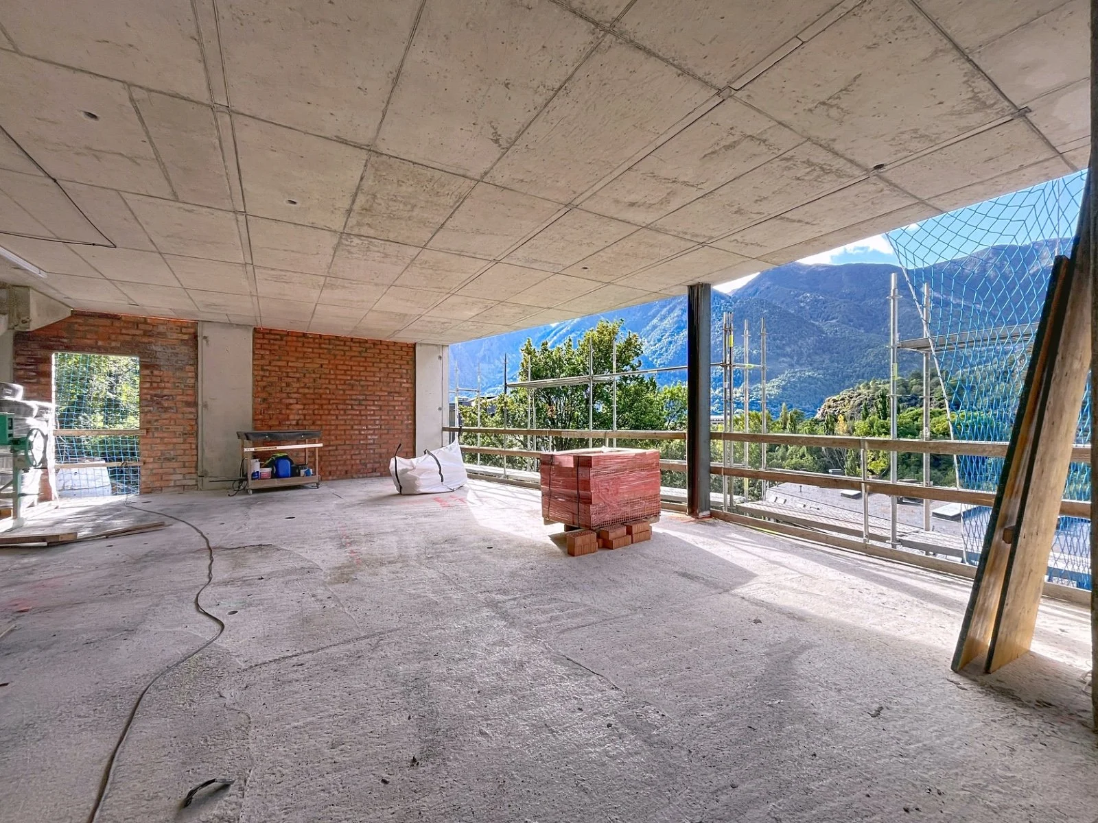Interiores de un edificio en construcción con vista a las montañas, piso de concreto y paredes de ladrillo, con materiales de construcción apilados y estructura de apoyo visible.